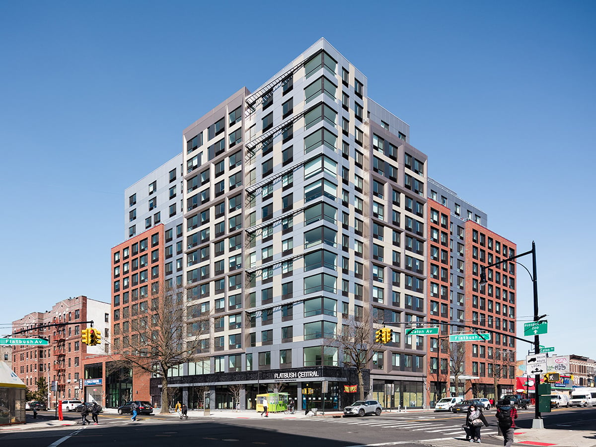 Street view of Caton Flats, Revitalized Flatbush Central Caribbean Marketplace. A modern mid-rise apartment building with large glass windows and red and grey exterior panels sits on a busy New York City street corner under a clear blue sky, with pedestrians and cars nearby. 