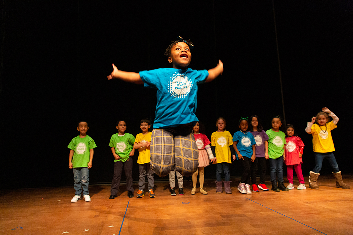 A young child in a blue shirt jumps energetically on stage with arms outstretched, while a group of children in colorful shirts stand in a line behind, watching. The background is dark and the stage is brightly lit.