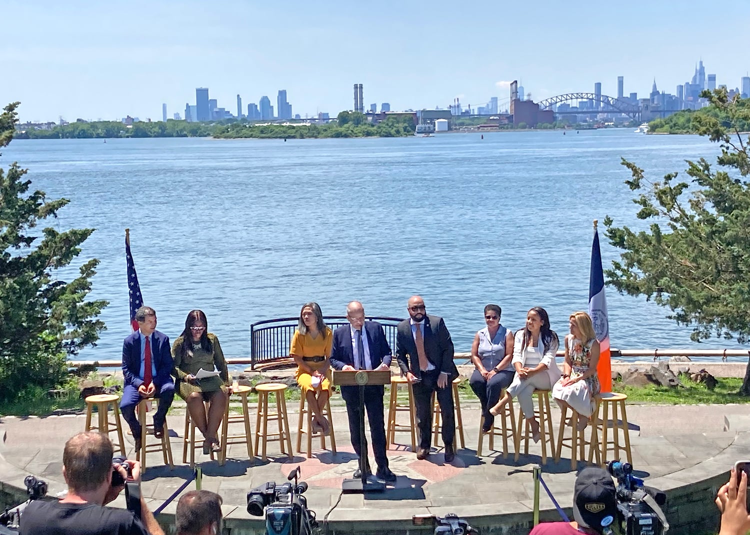 Group of eight community leaders sit on stools near a waterfront, with one person standing at a podium. Cameras film the release of Hunts Point Forward Vision, New York City skyline and bridge visible in the background, and two flags are displayed on each side.