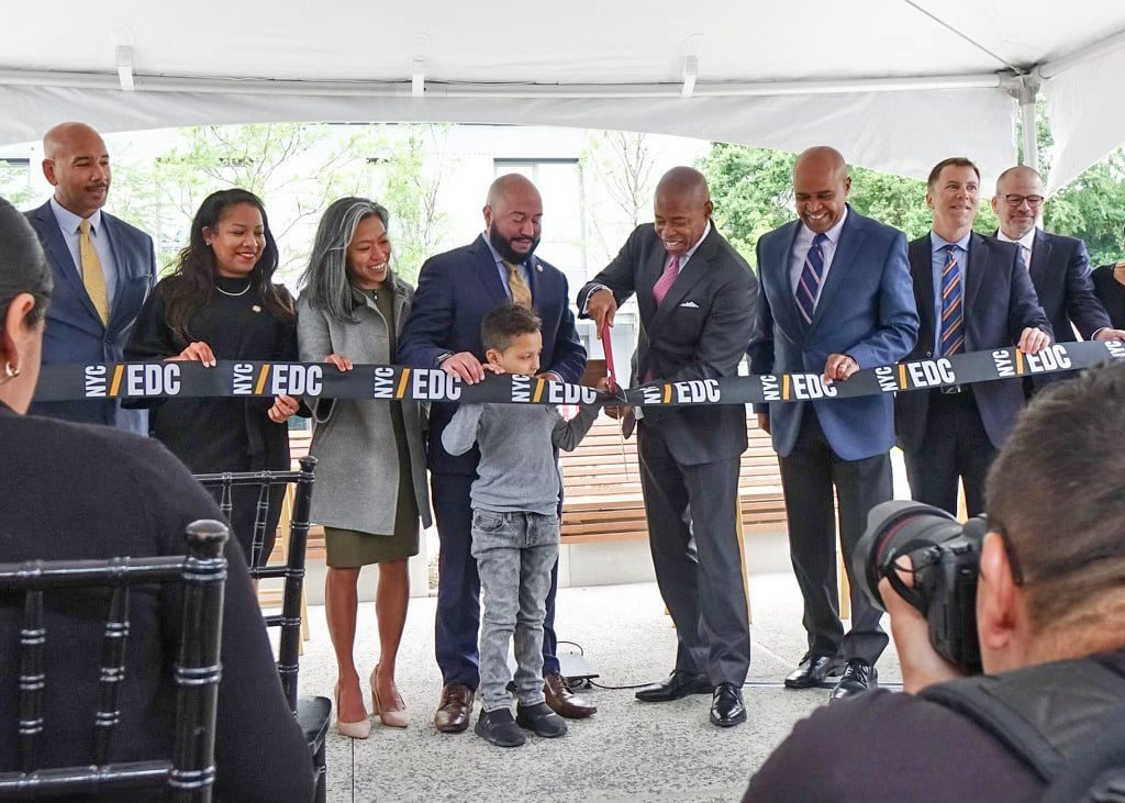 Mayor Adams along with a group of officials and a child, smile as they cut a ribbon labeled NYC/EDC to celebrate opening affordable homes on former site of Spofford Juvenile Detention Center. Photographers capture the moment at outdoor ceremony under a white tent.