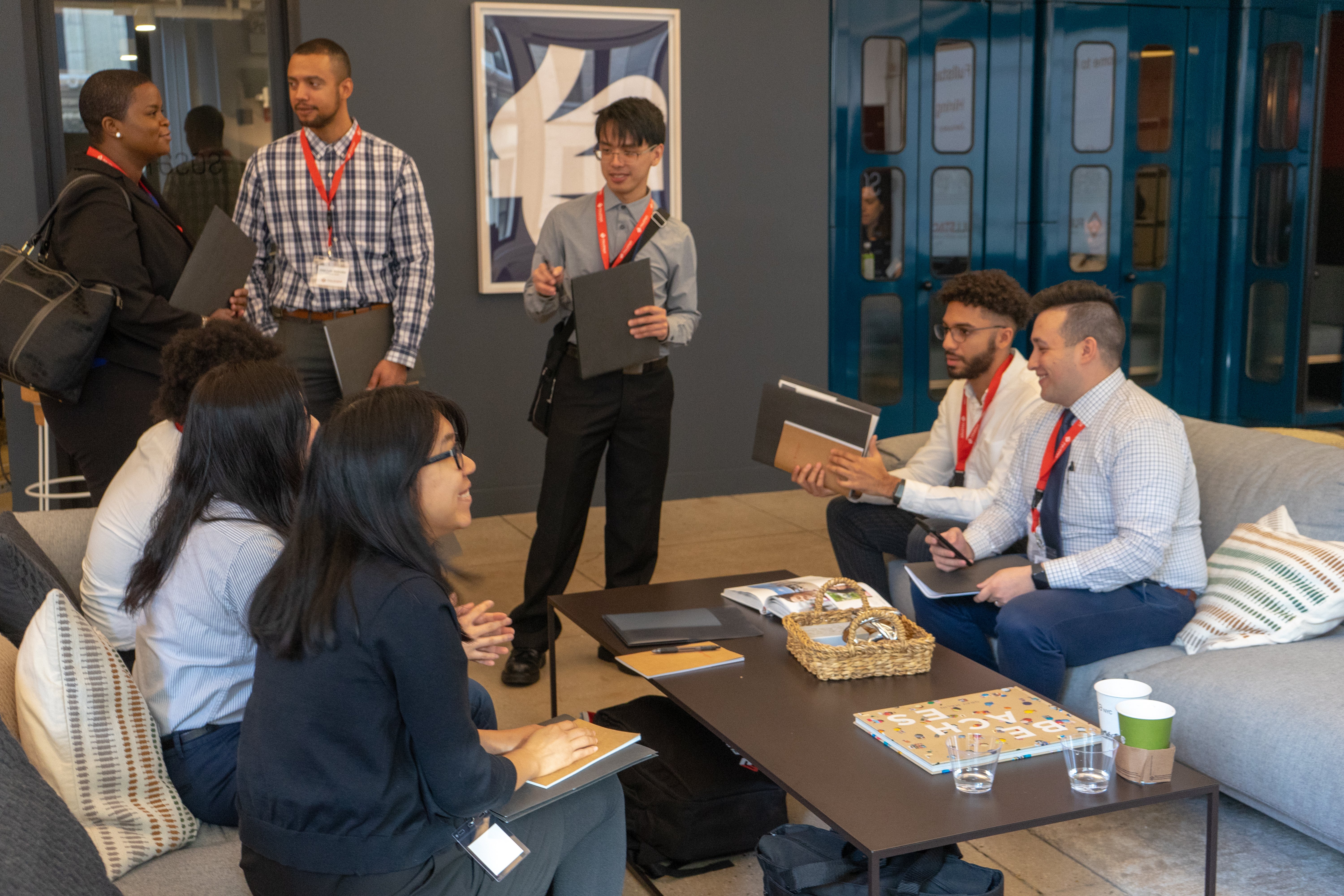 Fullstack Academy scholarship students at Cybersecurity Bootcamp wearing name badges and business attire sit and stand in a modern office lounge, talking and holding notebooks near a coffee table with snacks, drinks, and a board game.