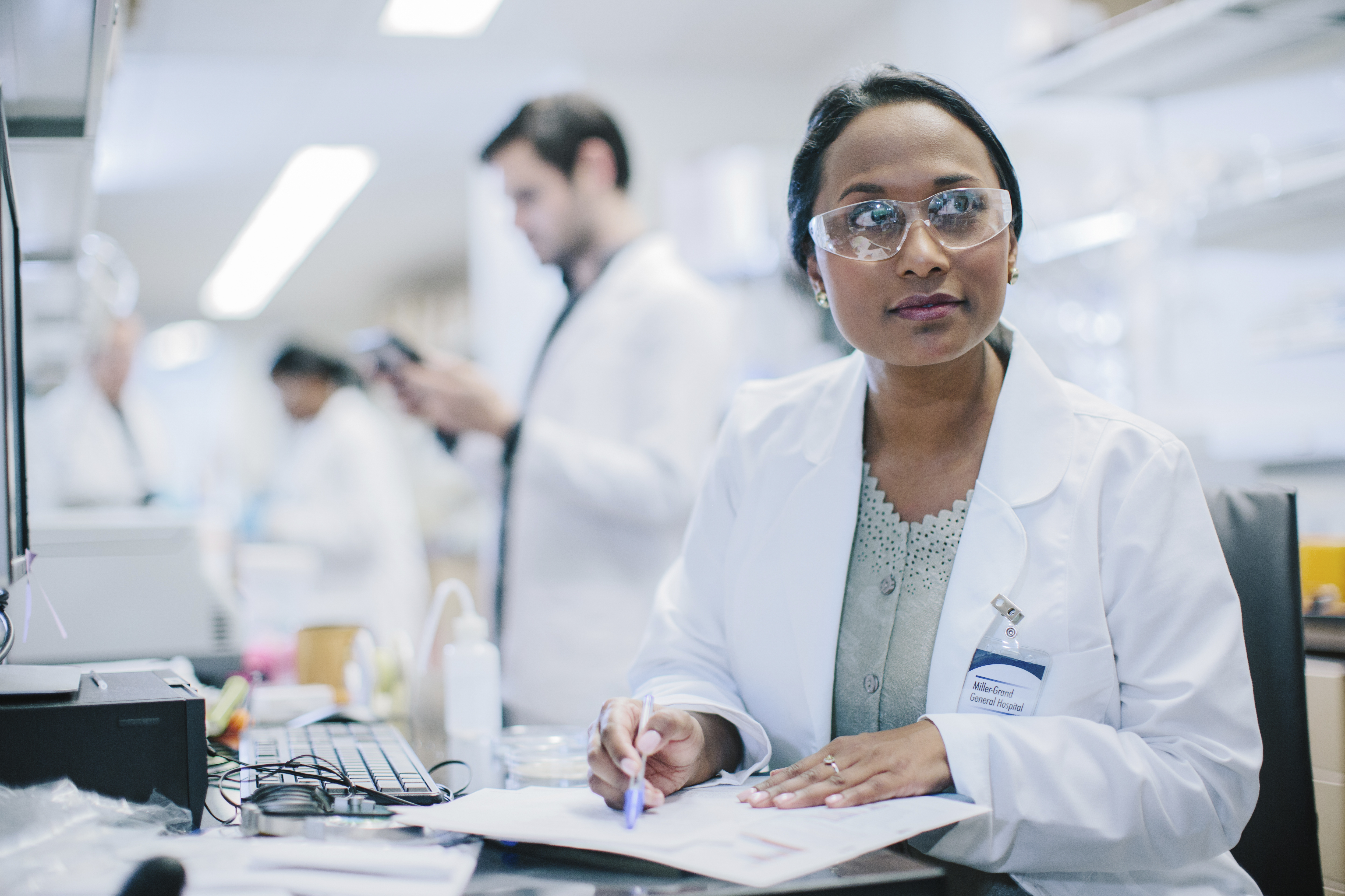 Female scientist wearing safety glasses and a lab coat sits at a lab desk, writing on a clipboard. Laboratory equipment and other scientists working in the background are visible.