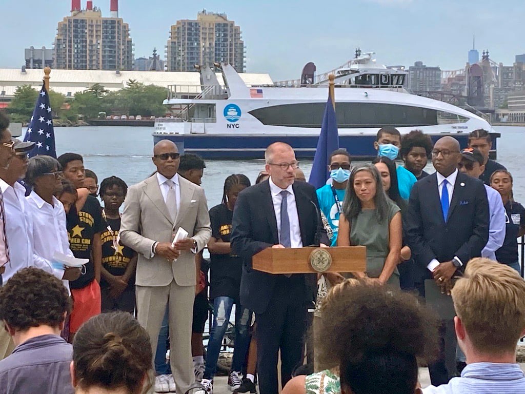 Group of people stand outdoors by an NYC Ferry waterfront, with a boat in the background. A suited Andrew Kimball speaks at a podium flanked by officials and attendees; flags are visible on either side, and Mayor Eric Adams is standing on the left of Kimball.