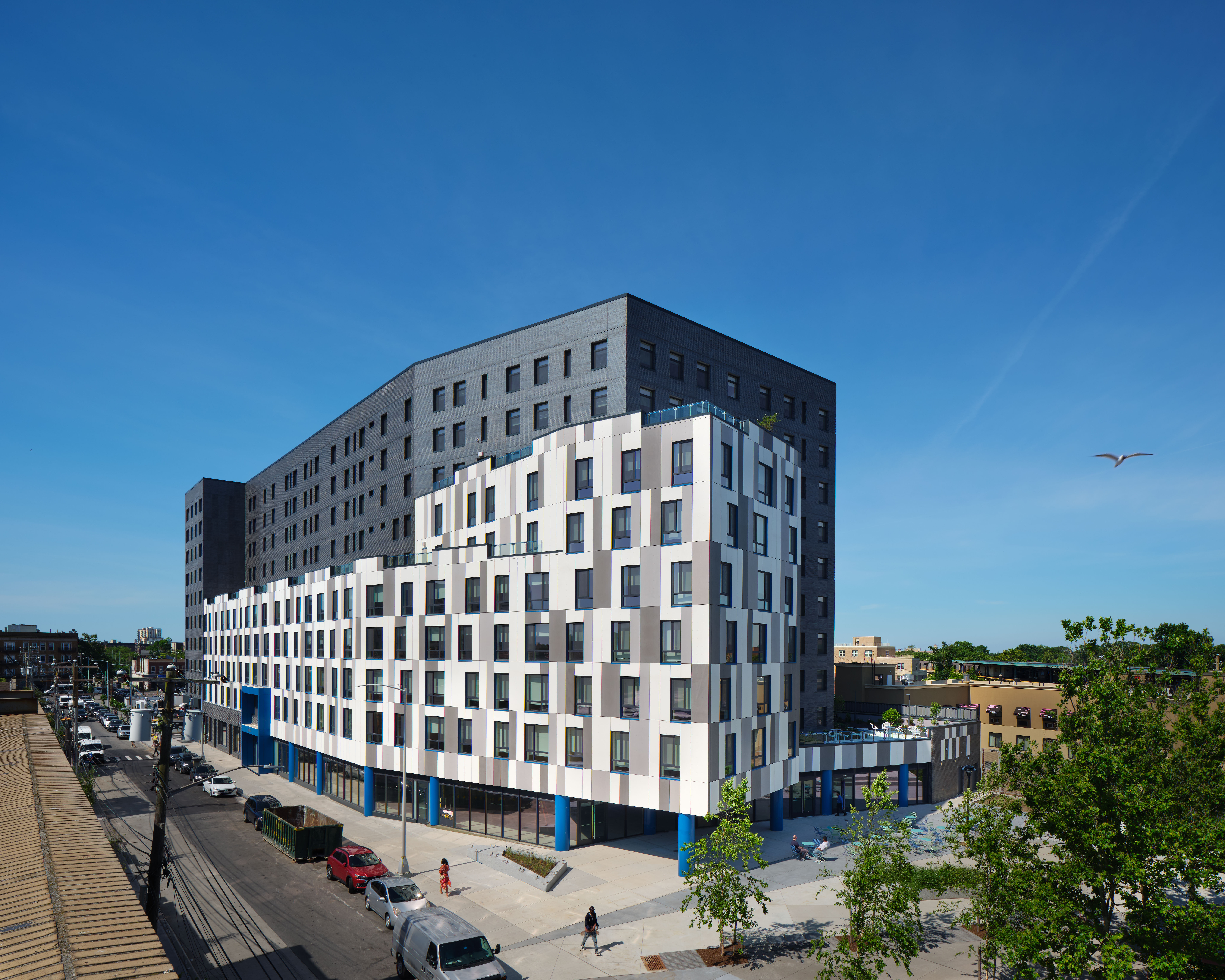 Image of Beach 21 shows a modern multi-story building with a black upper section and a white lower facade featuring irregular window patterns, located on a city street with cars, trees, and a clear blue sky above.