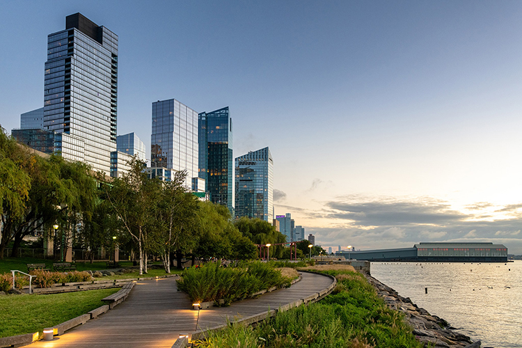 Image of Manhattan Waterfront Greenway shows a curved wooden pathway lined with greenery running along the waterfront at sunset, with modern glass skyscrapers and New York City buildings rising in the background.