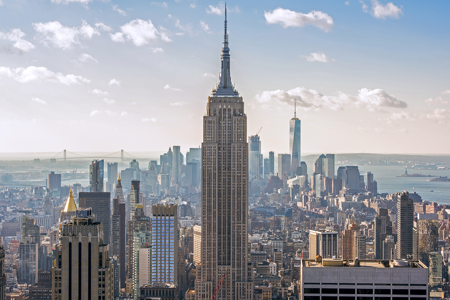 The Empire State Building stands tall in the center of a cityscape of Manhattan, New York, with other skyscrapers and distant bridges visible under a partly cloudy sky. Photo by Julienne Schaer.