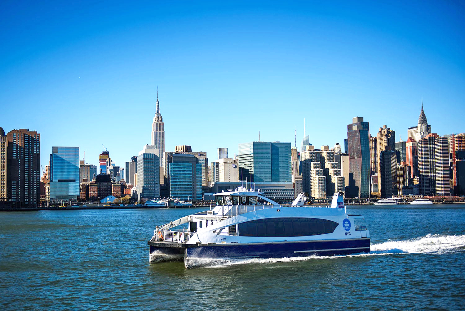 Modern NYC Ferry travels across the water with the New York City skyline, including the Empire State Building, visible under a clear blue sky in the background.