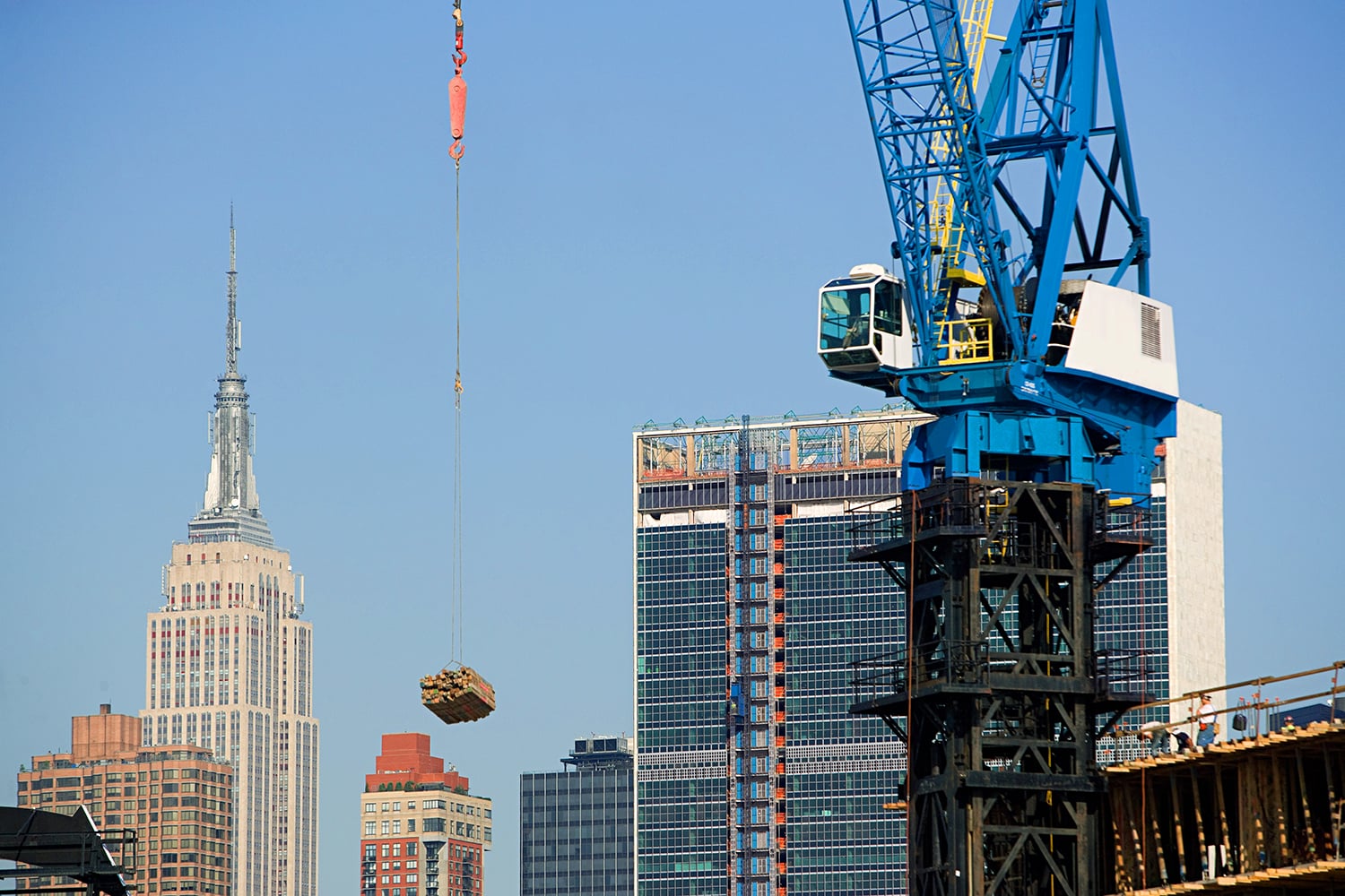 A construction crane lifts materials at a building site in NYC, with the Empire State Building and other skyscrapers visible in the background under a clear blue sky.