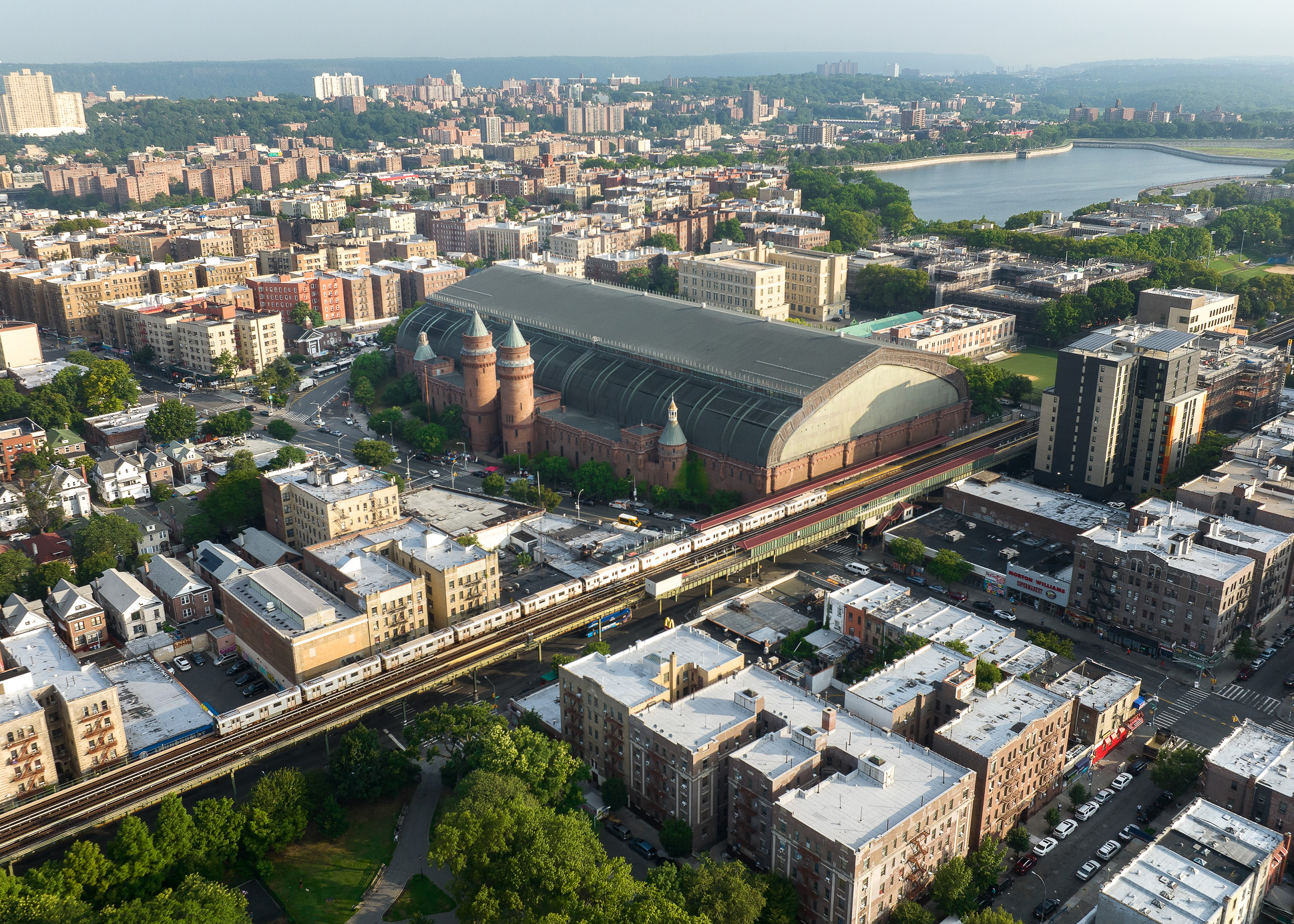 Aerial rendering of Kingsbridge Armory, a large brick building with a curved glass roof, surrounded by residential and commercial buildings. A train is visible on tracks in the foreground, with greenery and a body of water in the background.