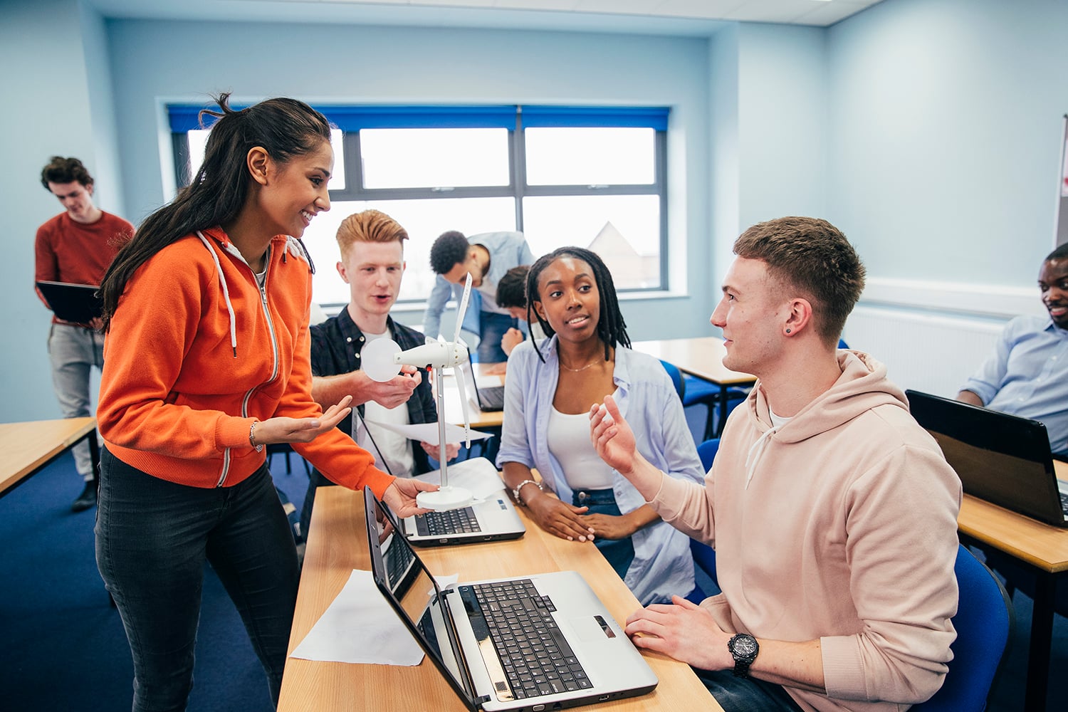 A group of college students in a classroom are collaborating, smiling, and discussing while using laptops. One student stands, holding a 3D-printed wind turbine model, engaging with seated classmates in a bright, modern room.