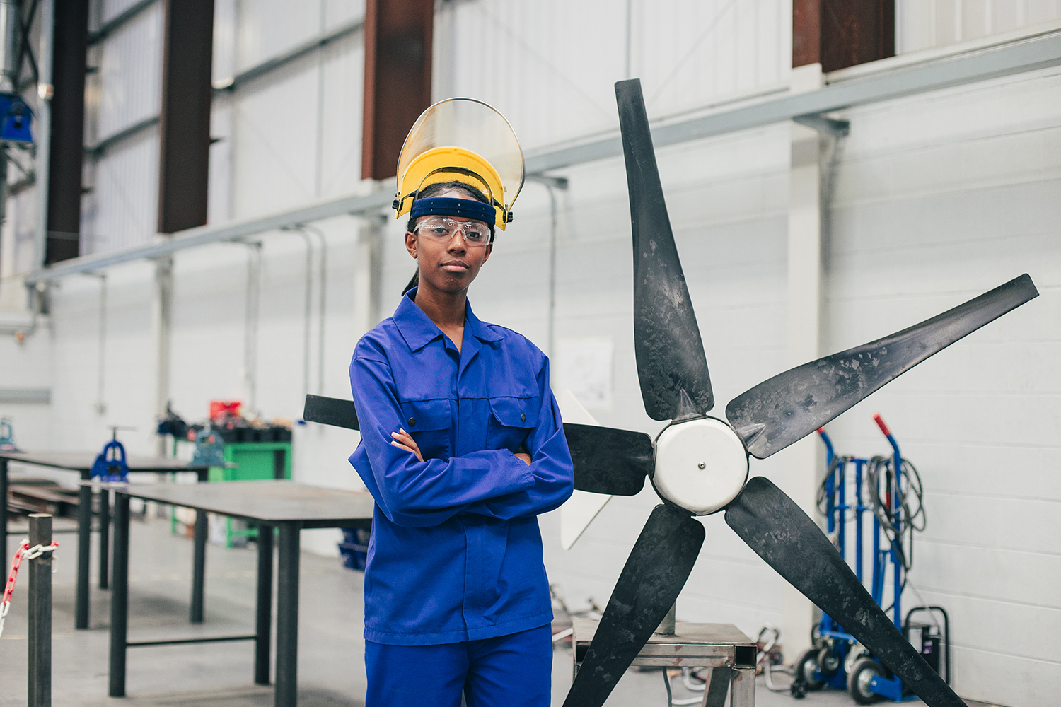 A woman in blue work attire and safety gear stands with arms crossed in an industrial setting, next to a large fan blade. The background shows workbenches and equipment, indicating a workshop or manufacturing environment.