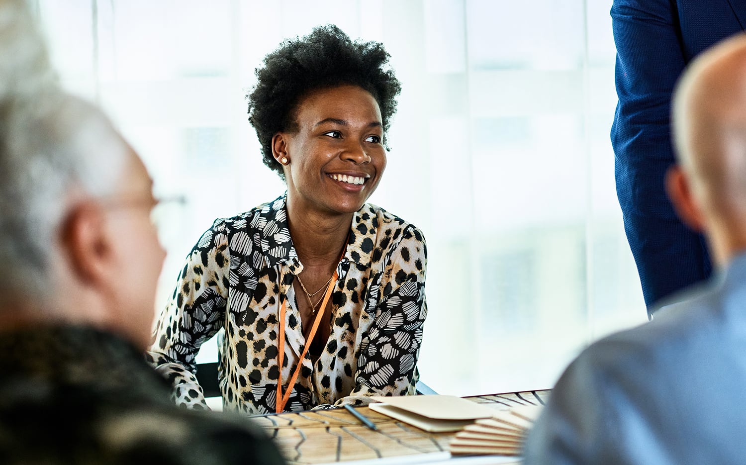 A smiling mid-adult Black woman with short curly hair and a patterned blouse sits at a table with others in a bright, modern setting. She appears engaged in conversation, with notepads and papers in front of her.