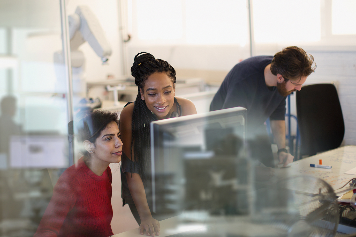 Three people in an office are engaged with computer work. Two are focused on a monitor, while the third is writing on a whiteboard in the background. The atmosphere is collaborative and professional.