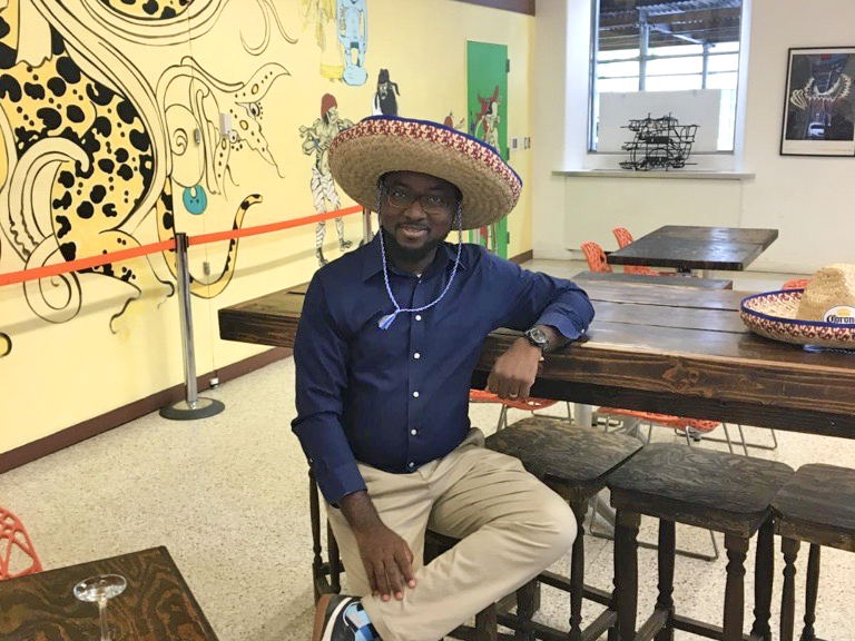 Sakiru Okeowo wearing a large straw sombrero sits casually at a wooden table in a vibrant room with colorful murals and a rope barrier. He is dressed in a blue shirt and beige pants, with a relaxed expression.