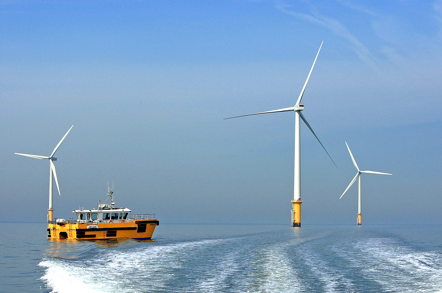 A yellow workboat sails near three large offshore wind turbines in calm, blue water under a clear sky, leaving a white wake behind.