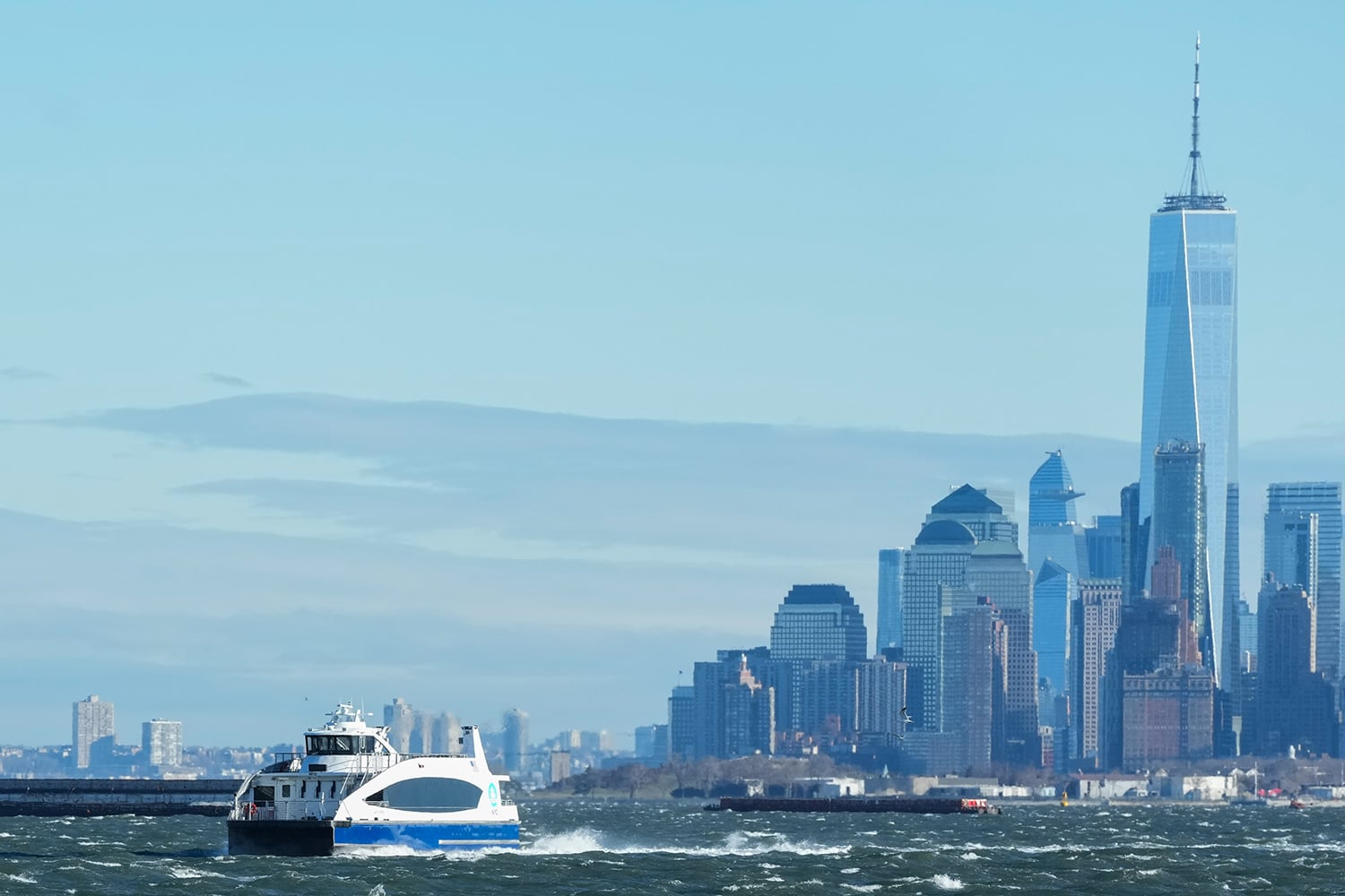 A white and blue NYC Ferry travels across choppy water with the New York City skyline, including the tall One World Trade Center, visible in the background under a clear sky.
