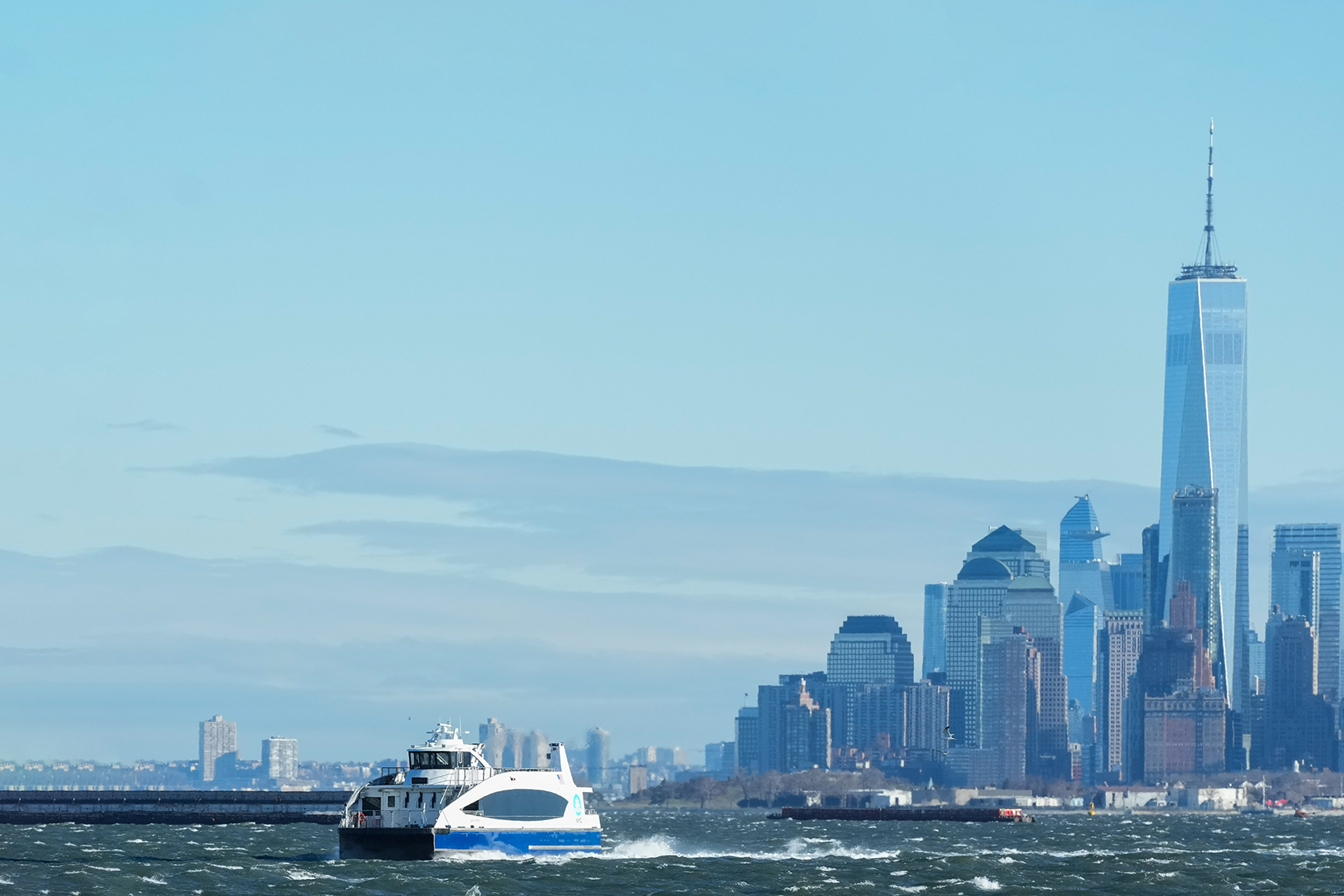 White and blue NYC Ferry travels across choppy water with the New York City skyline, including the One World Trade Center, visible in the background under a clear sky.