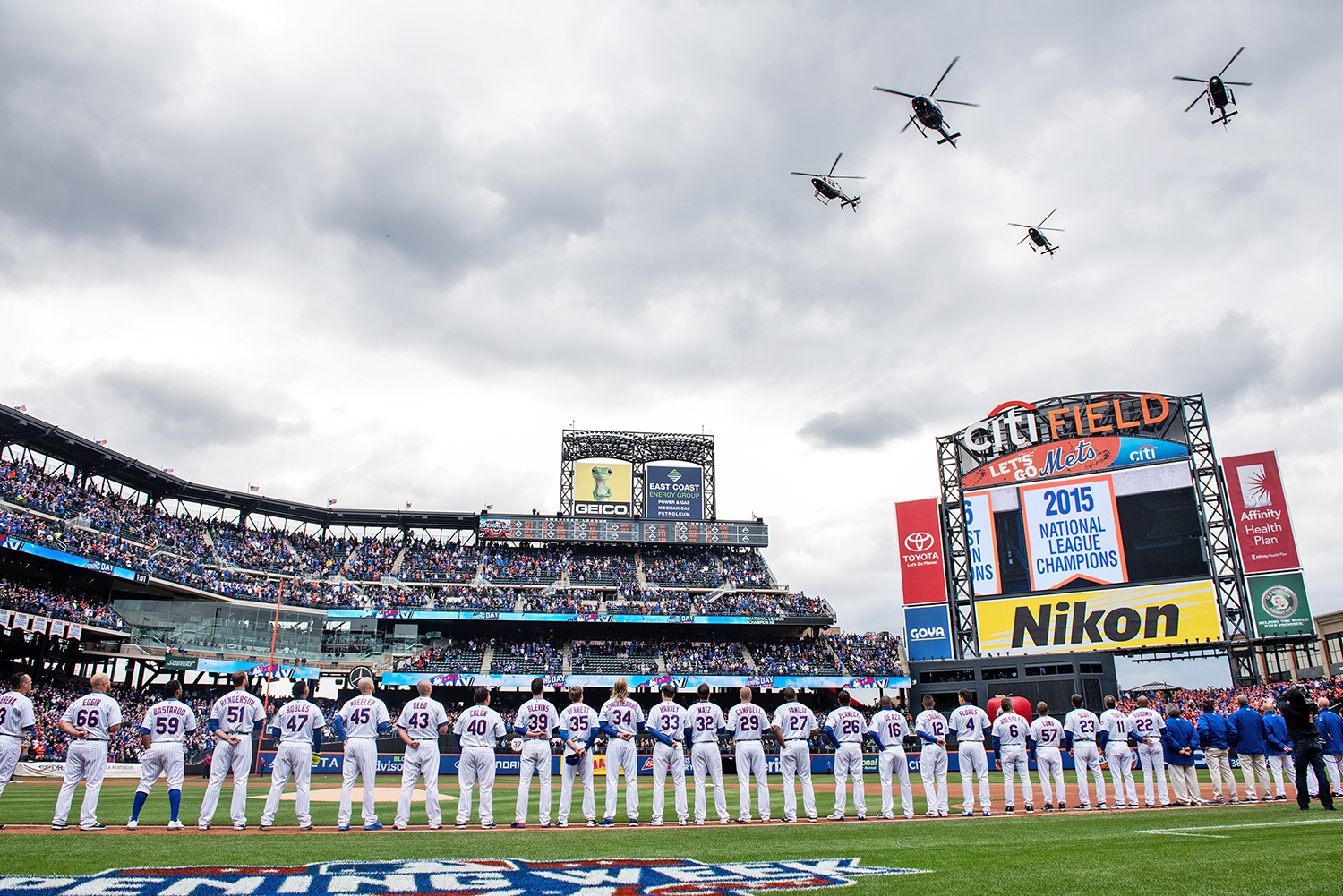 New York Mets players in uniform line up on the field at Citi Field, with a crowd in the stands and four helicopters flying overhead. A scoreboard displays “2015 National League Champions” for the New York Mets.