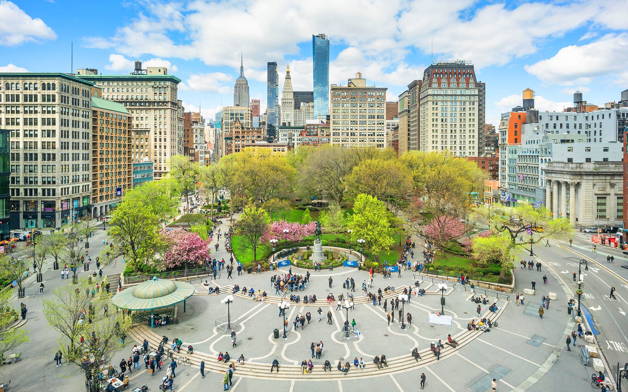 Aerial view of New York's Union Square, a lively urban park surrounded by tall buildings under a blue sky with clouds. The park features lush green trees, blooming pink flowers, a central fountain, and numerous people walking and sitting.