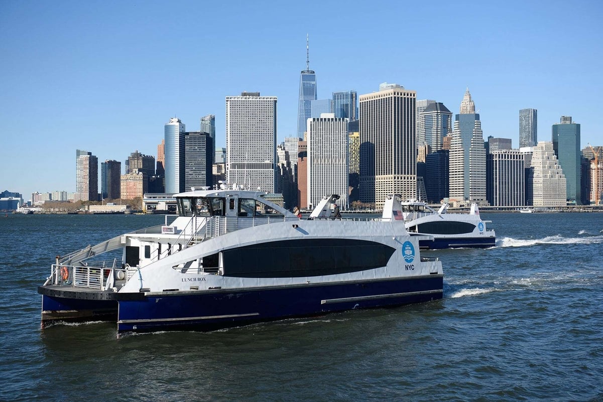 Two NYC ferry boats travel on the water with the Manhattan skyline, including One World Trade Center, visible in the background on a clear day.