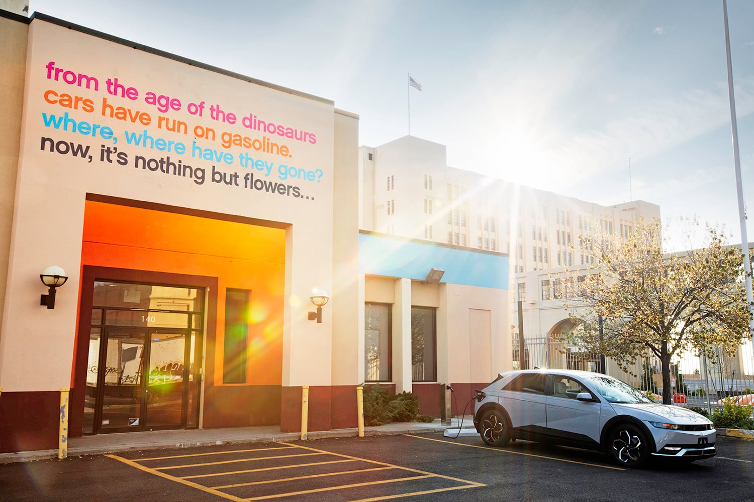 A silver electric car charging while parked at a curb outside a building with text on the wall referencing dinosaurs, gasoline, and flowers. The sun shines brightly, casting long shadows, and a large government building is visible in the background. The charging port is by itselectric, a Brooklyn-based electric vehicle curbside charging company.