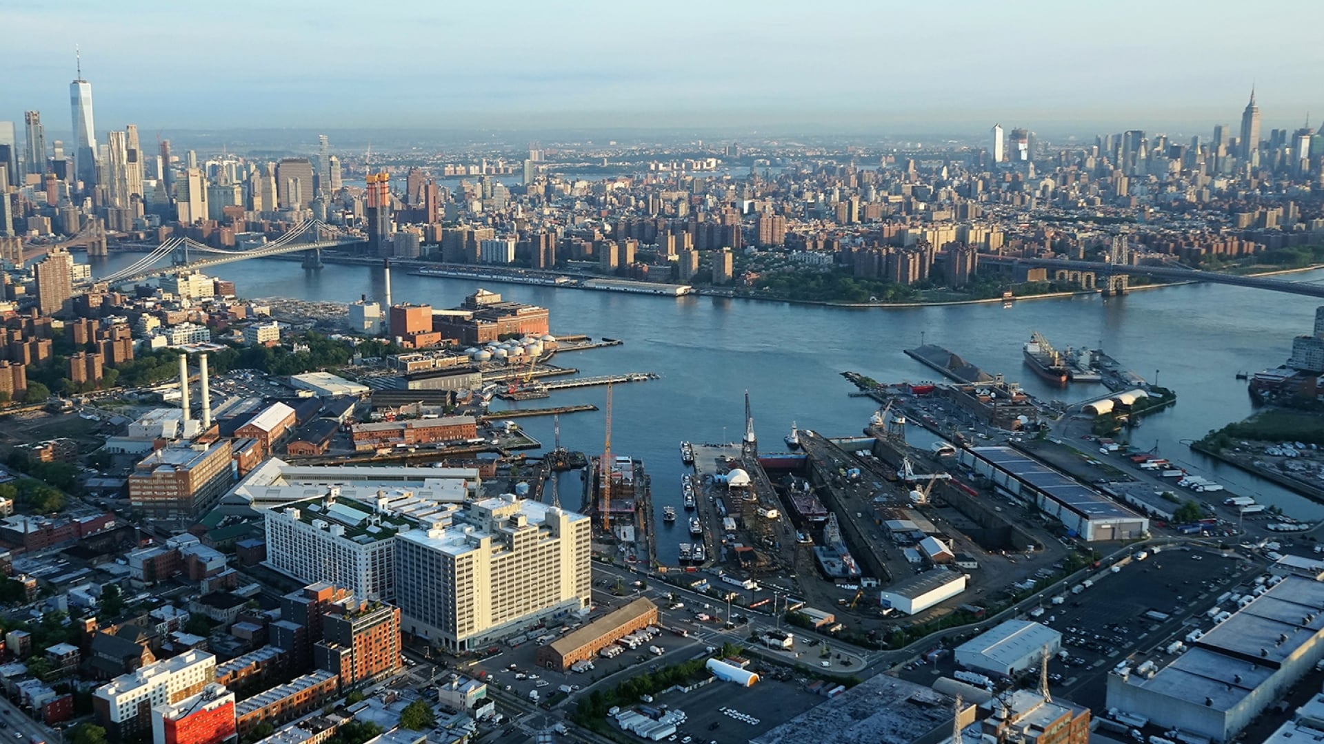 Aerial view of Brooklyn Navy Yard with piers, warehouses, and streets in the foreground, and the East River separating Brooklyn from the Manhattan skyline in the background, including several tall skyscrapers. Photo by Jerry Trudell.