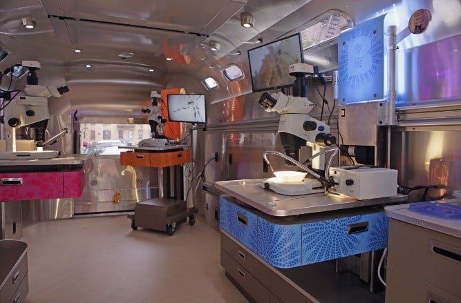 The interior of BioBus, a modern mobile laboratory with workstations equipped with microscopes, monitors, and bright, colorful panels. There are screens, lights, and scientific equipment on countertops throughout the space.