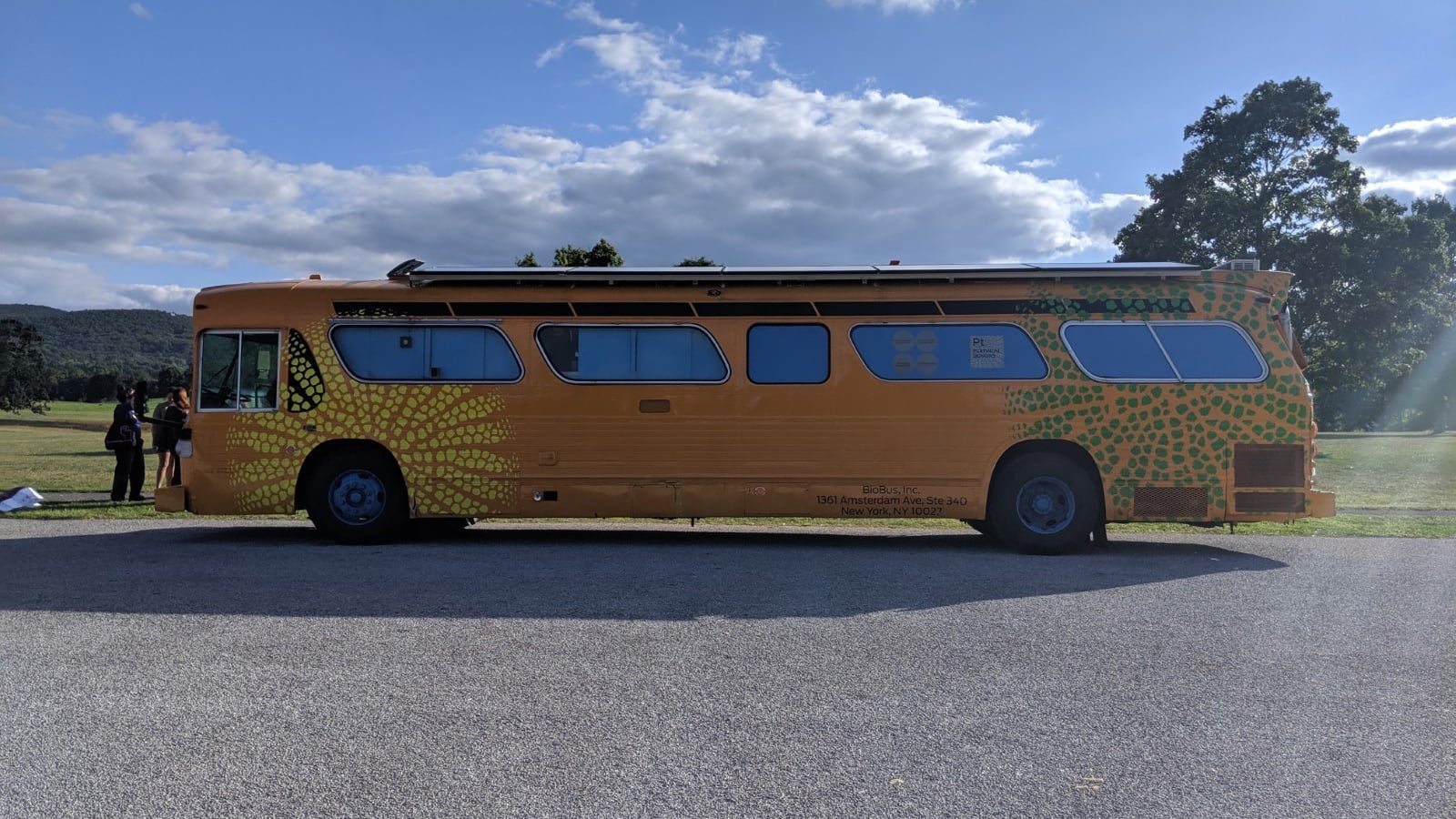 Large yellow BioBus with green circular patterns is parked on a paved area beside a grassy field with trees and hills in the background under a partly cloudy sky. A few people stand near the bus.