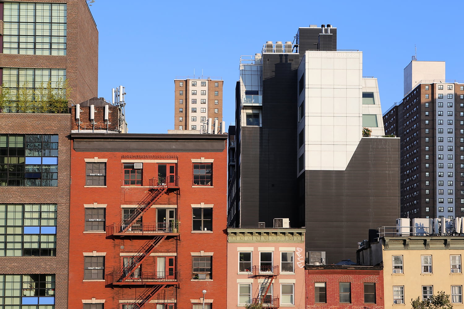 A row of urban buildings, including red-brick apartments with fire escapes and modern high-rises, stands against a clear blue sky in Meatpacking District, Manhattan.