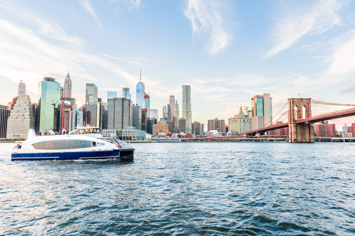 View of NYC Ferry with Lower Manhattan skyline and Brooklyn Bridge in the background at sunset.