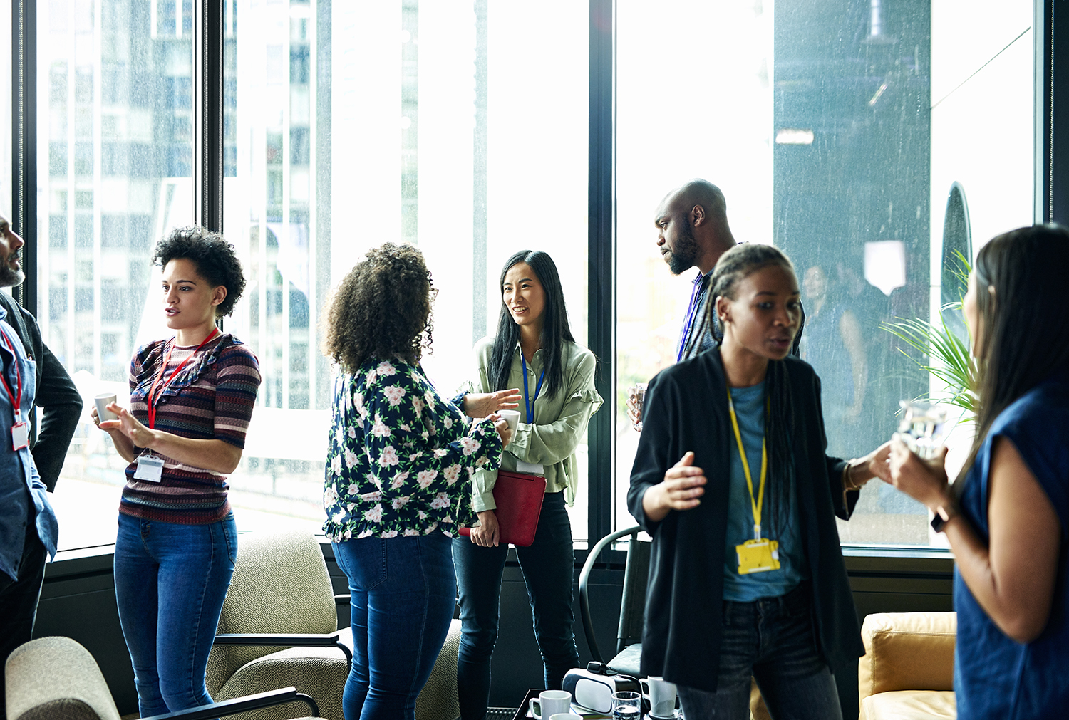 A group of people participating in NYCEDC Fellowship Program stand in a brightly lit room with large windows, engaging in conversation. They are casually dressed and appear to be in an office or conference setting. Coffee cups are on a table nearby.