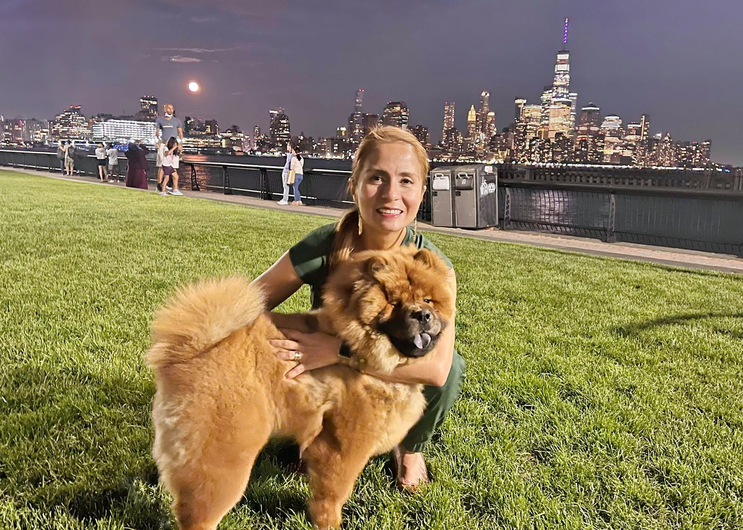 Diana Franco smiles while hugging her fluffy brown dog on a grassy area at night. The city skyline, illuminated, is in the background across a body of water, with a full moon visible in the sky.