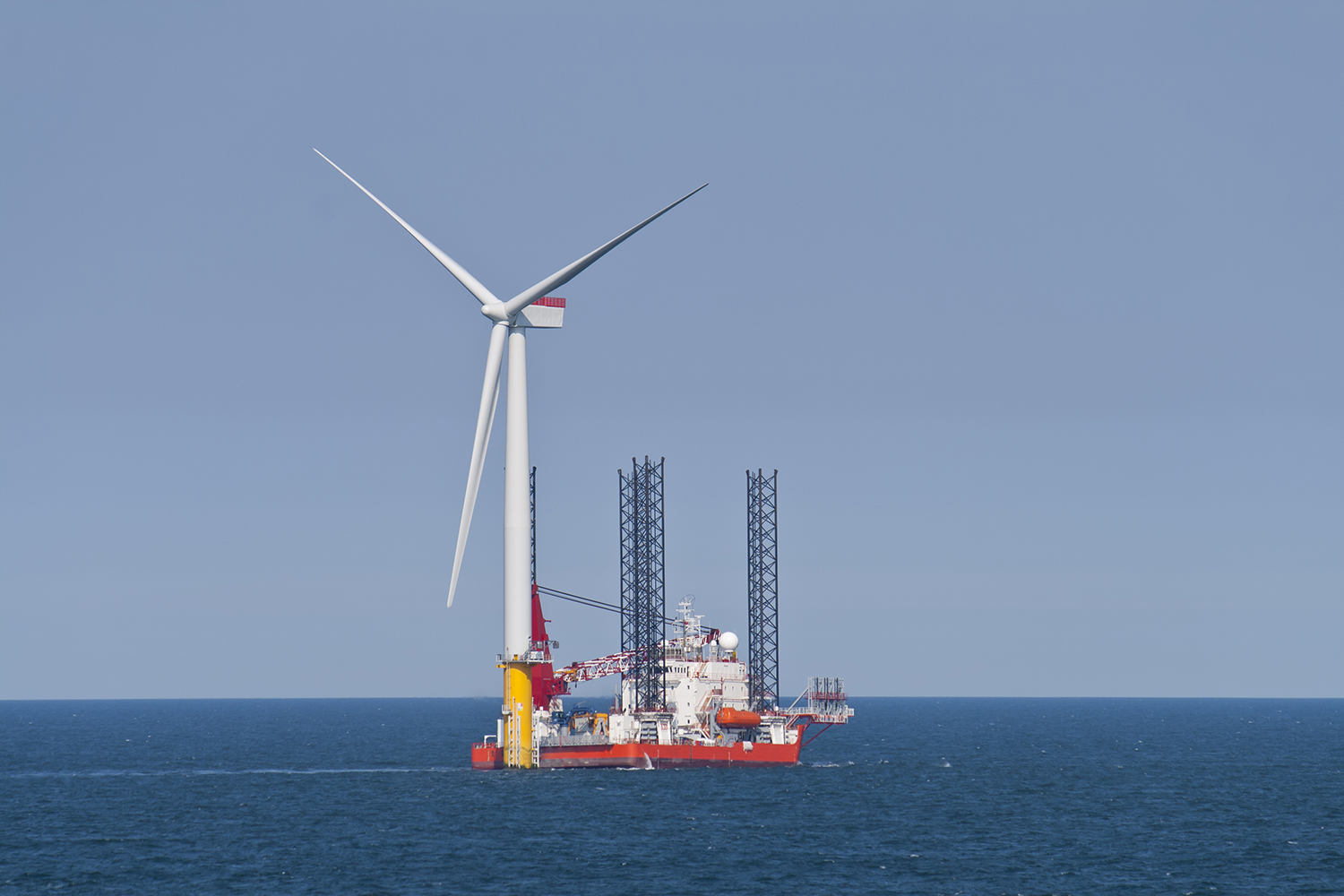A large wind turbine under construction stands in the ocean next to a red and white offshore service vessel equipped with cranes and tall support legs, under a clear blue sky.
