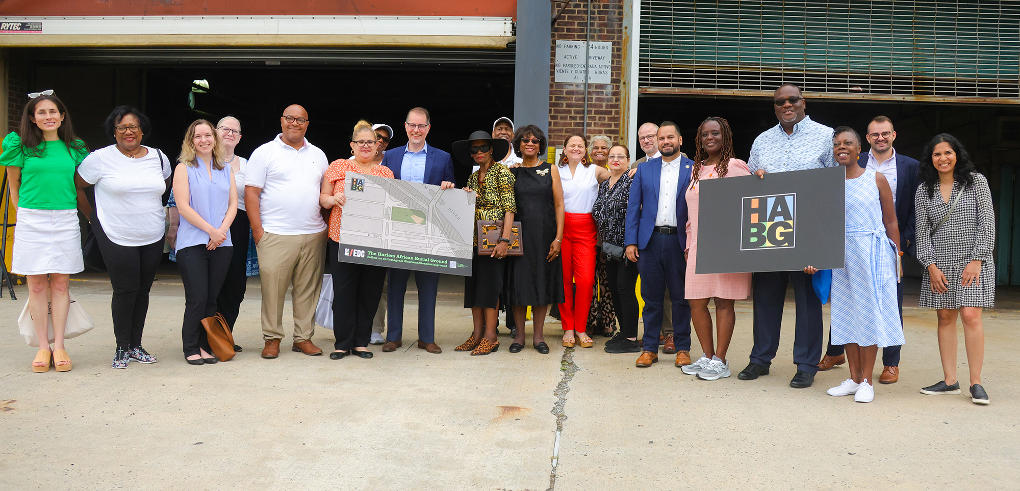 Diverse group of community leaders stands in front of a partially open garage, some holding signage and a map, smiling for a group photo on a sunny day. They are there to announce the next phase of archaeological work at the Harlem African Burial Ground.