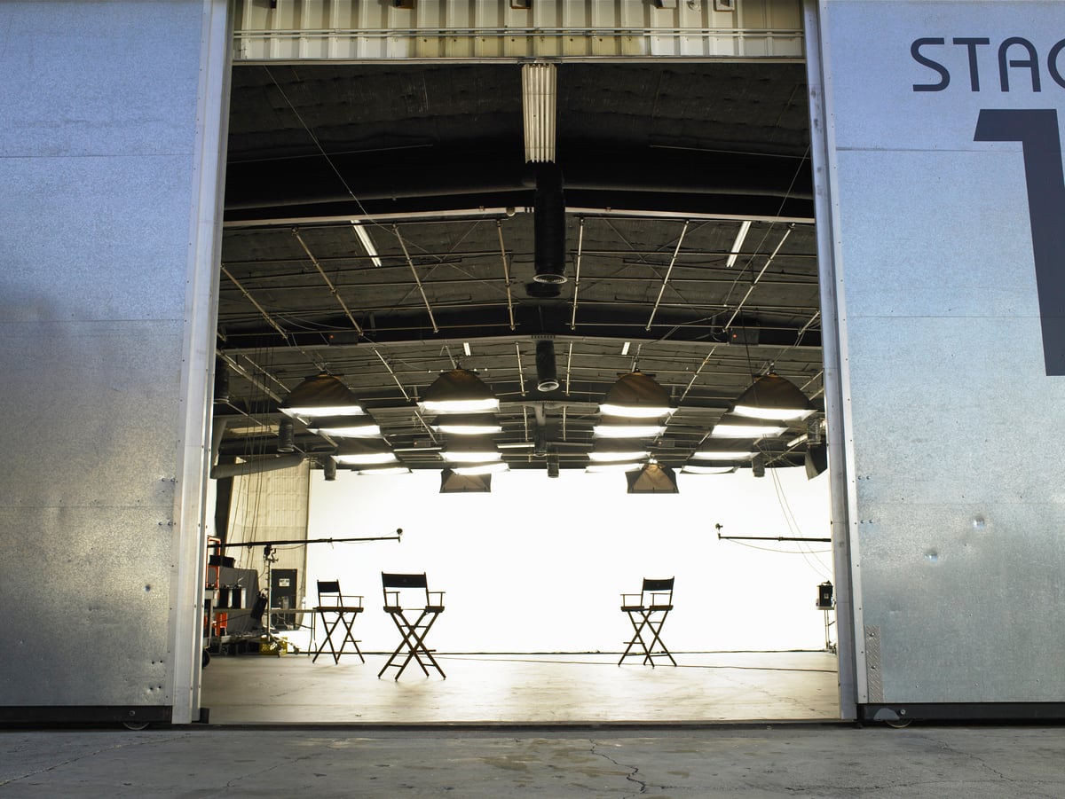 An open studio stage door reveals a brightly lit interior with four empty director’s chairs arranged in a row, film equipment visible to the side, and a white backdrop in the background.