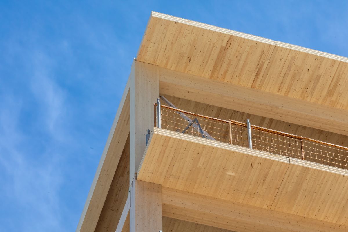 Close-up of a modern wooden building structure with visible beams and a balcony railing, set against a bright blue sky.