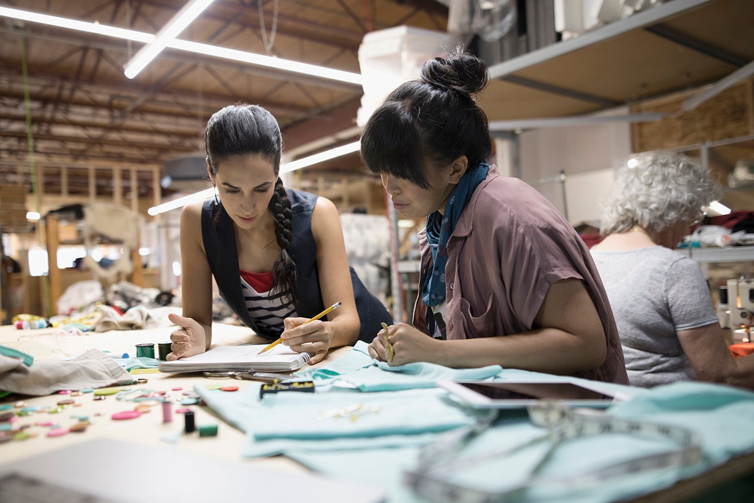 Two women fashion designers work together at a table covered with fabric, thread, and sewing supplies in a workshop, focusing on a sketchbook. Another woman in the background sews at a machine. The space is busy and creative.