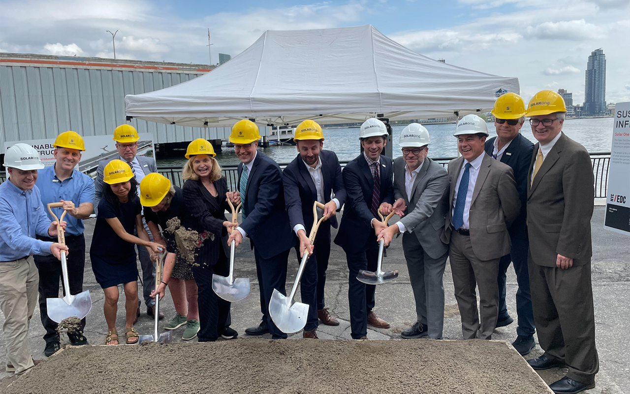 Group of people from NYCEDC and Solar One in business attire and yellow hard hats at Solar One Environmental Education Center Groundbreaking. They are shoveling dirt and standing in front of a white tent near a waterfront.