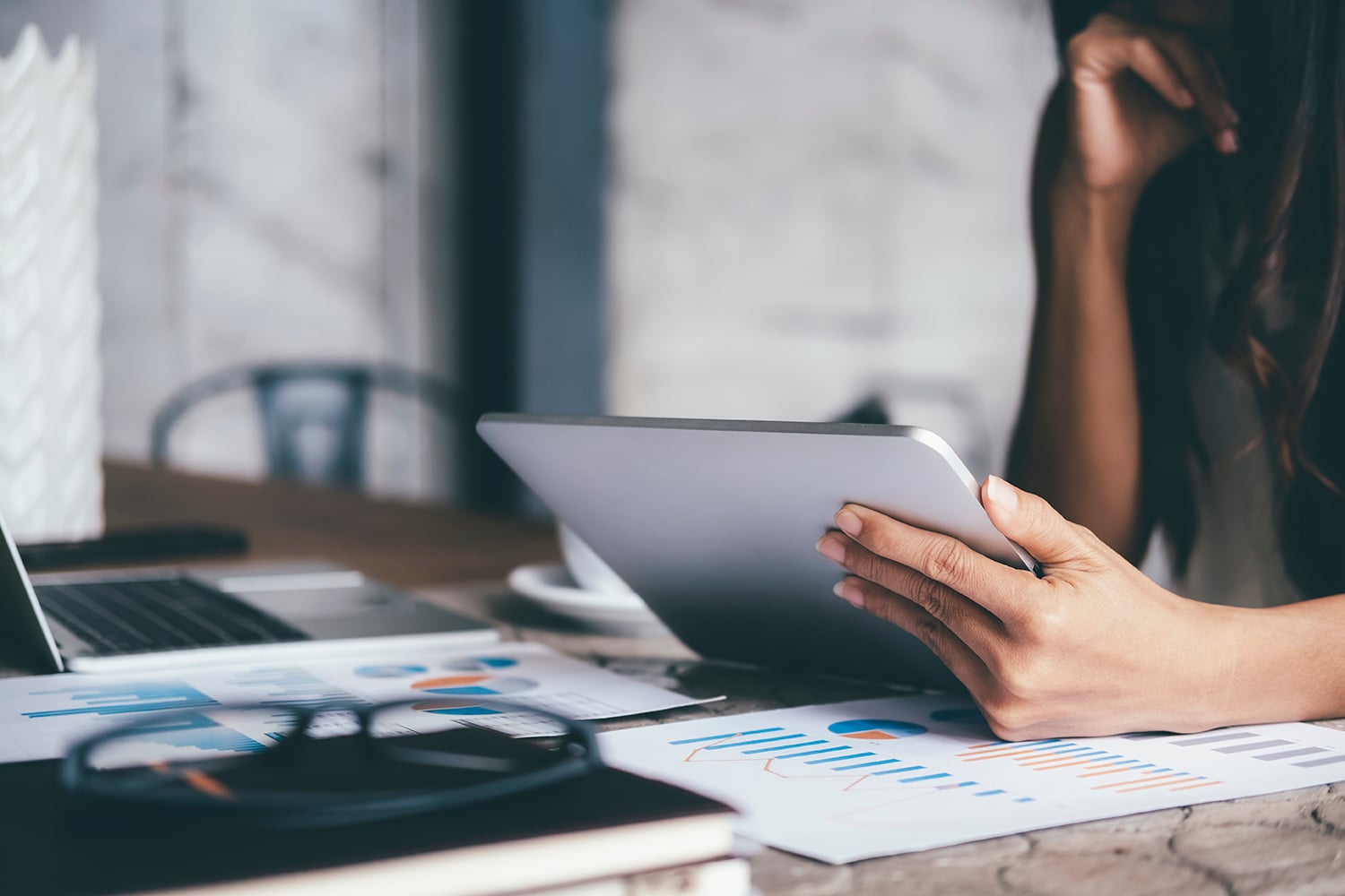 Businesswoman holding a tablet sits at a desk with business charts, a laptop, and glasses, analyzing investment marketing data in a modern office environment.