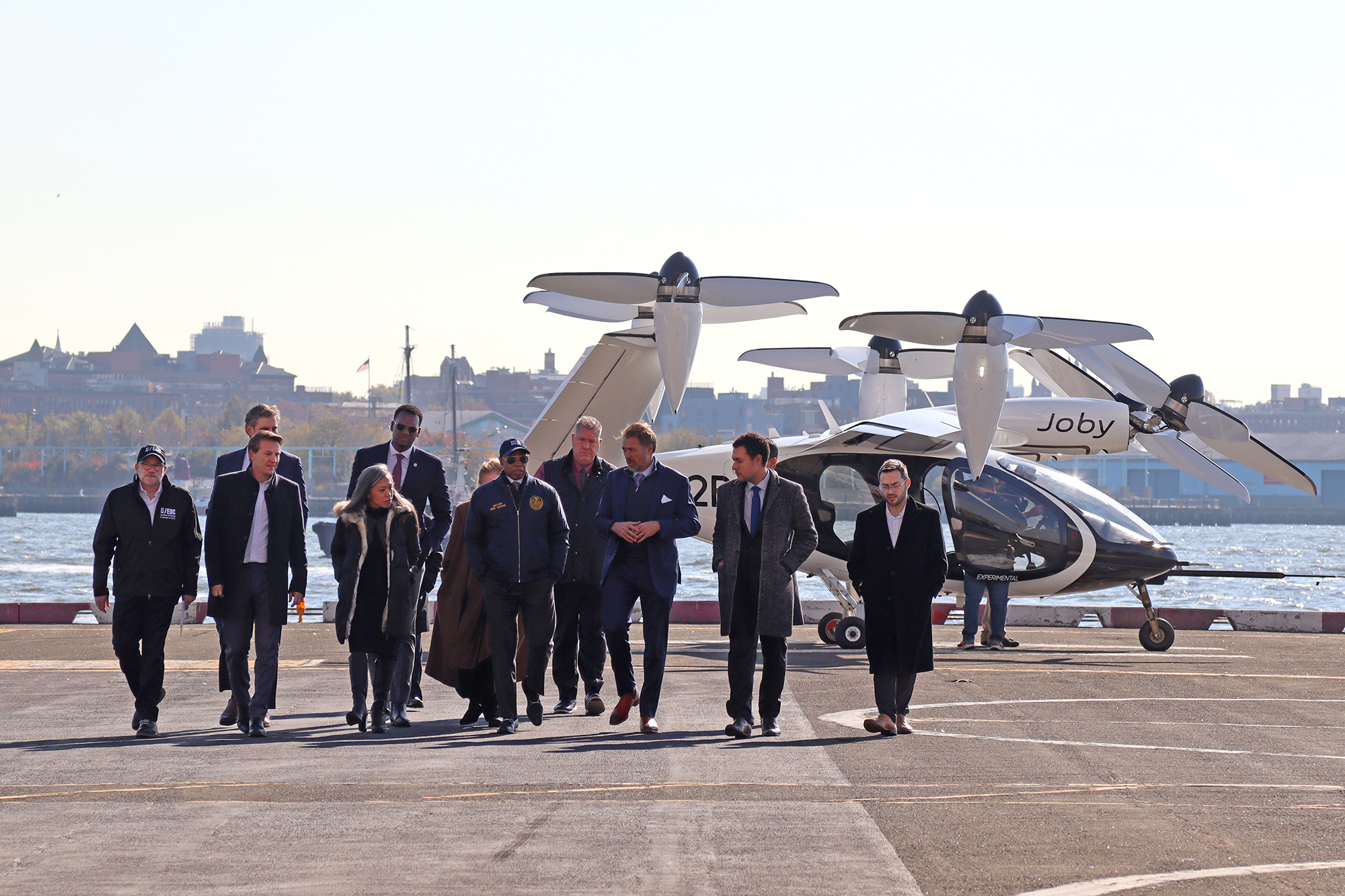 NYC Mayor Adams and leaders in business and official attire walking in front of an electric vertical take-off and landing (eVTOL) aircraft, with a river and the New York Skyline in the background.