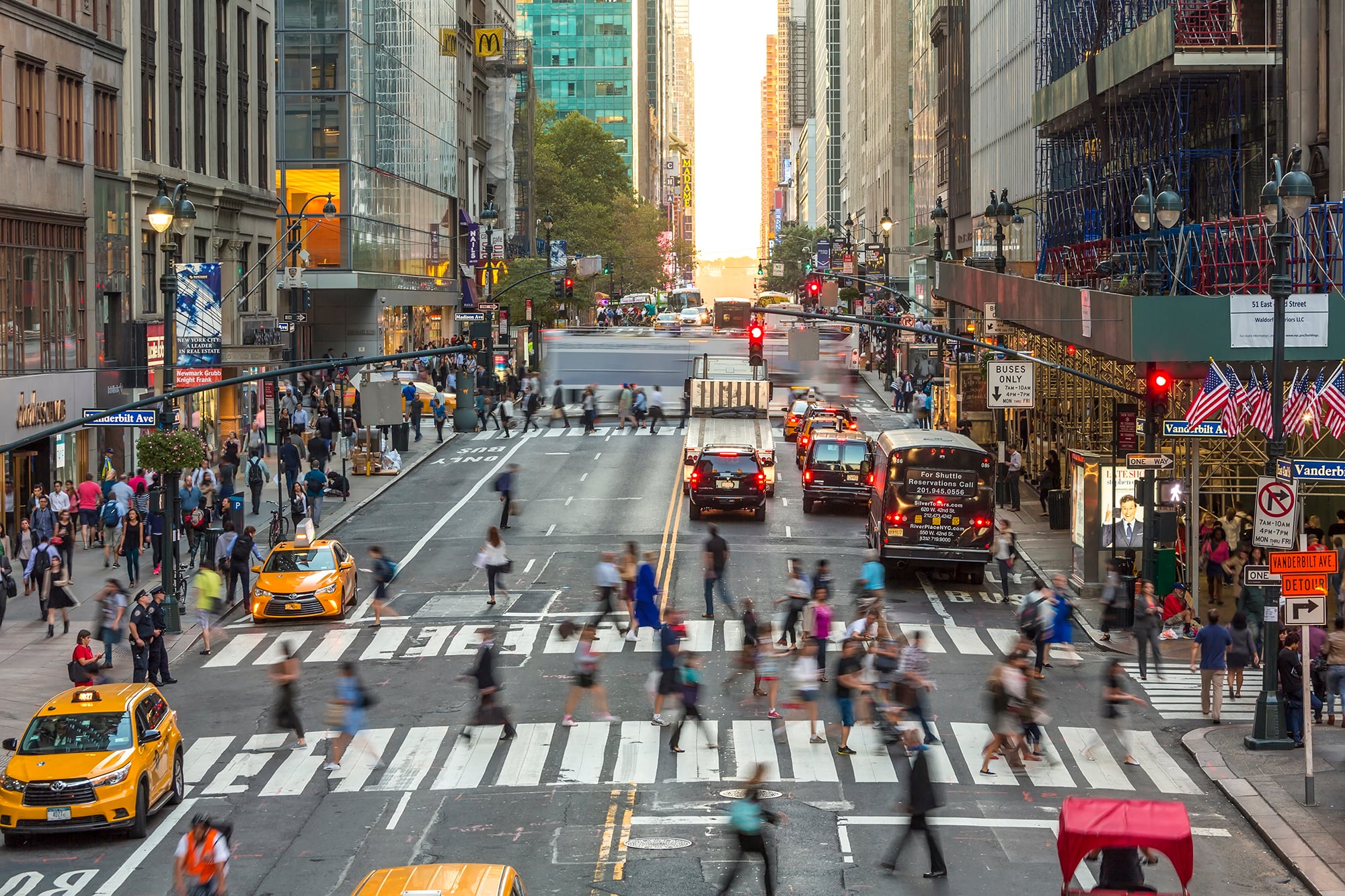 A busy 42nd Street in Midtown Manhattan, New York City, with many people crossing at crosswalks, yellow taxis, cars, buses, and tall buildings on both sides. Traffic lights and storefronts are visible, and the scene appears lively and bustling.
