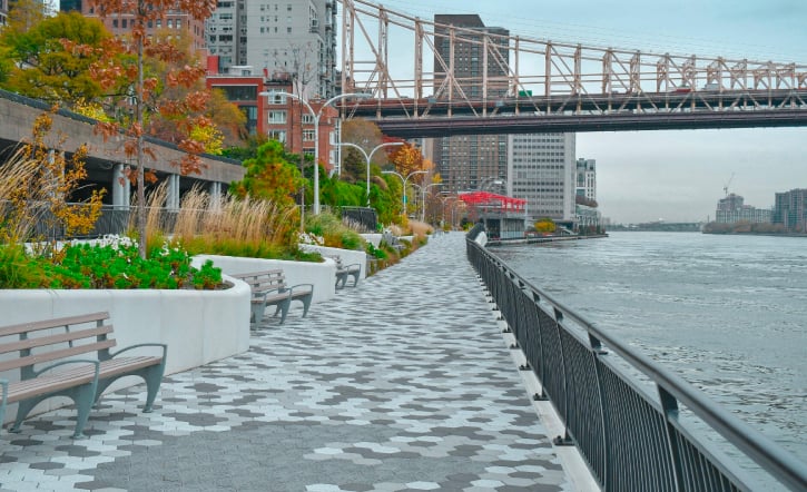 Rendering of East Midtown Greenway and Phase 2B of Andrew Haswell Green Park shows a paved riverside walkway with benches and greenery that run alongside a body of water, with Brooklyn Bridge and Manhattan buildings in the background. Trees with autumn foliage line the path.
