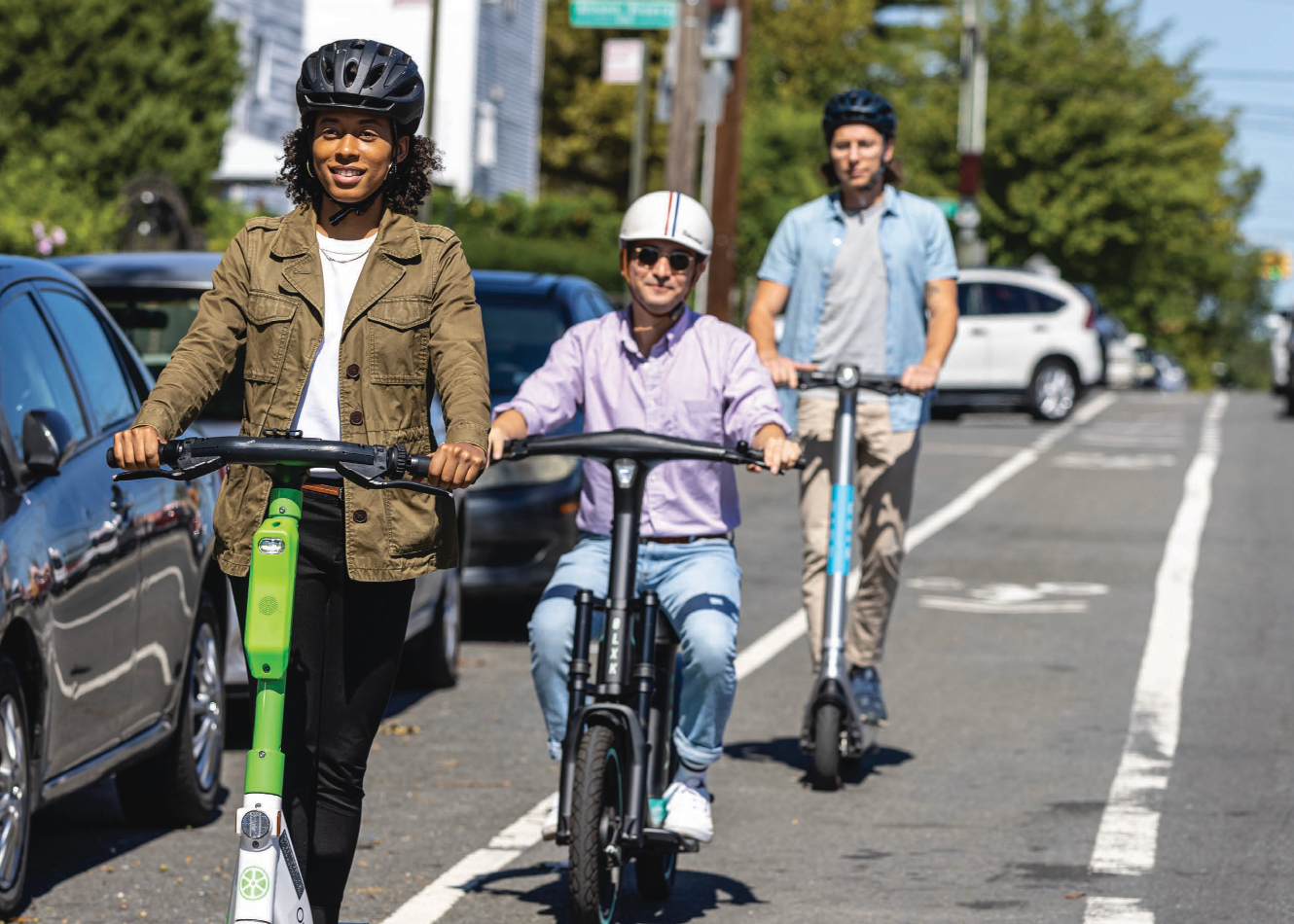Two men and one woman riding e-bikes and scooters in an NYC bike lane with helmets on.
