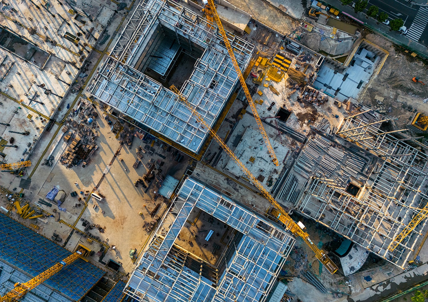 Aerial view of a construction site with multiple buildings in progress. Steel frameworks and construction cranes are visible, along with scattered materials and equipment. The site is busy with activity, under clear weather conditions.