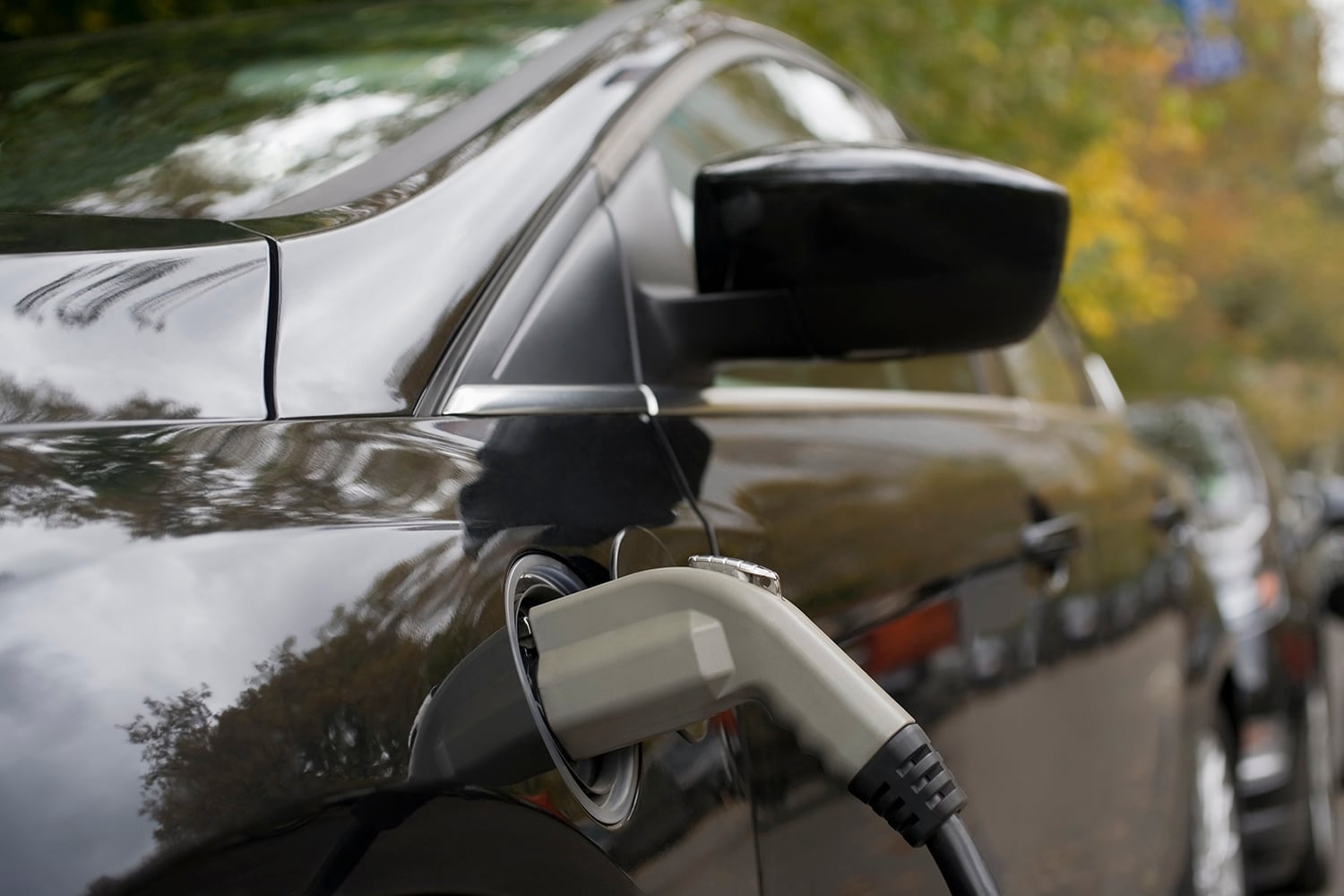 Close-up of an electric car being charged, with a charging cable plugged into the vehicle. The car’s glossy black exterior reflects trees and greenery in the background.