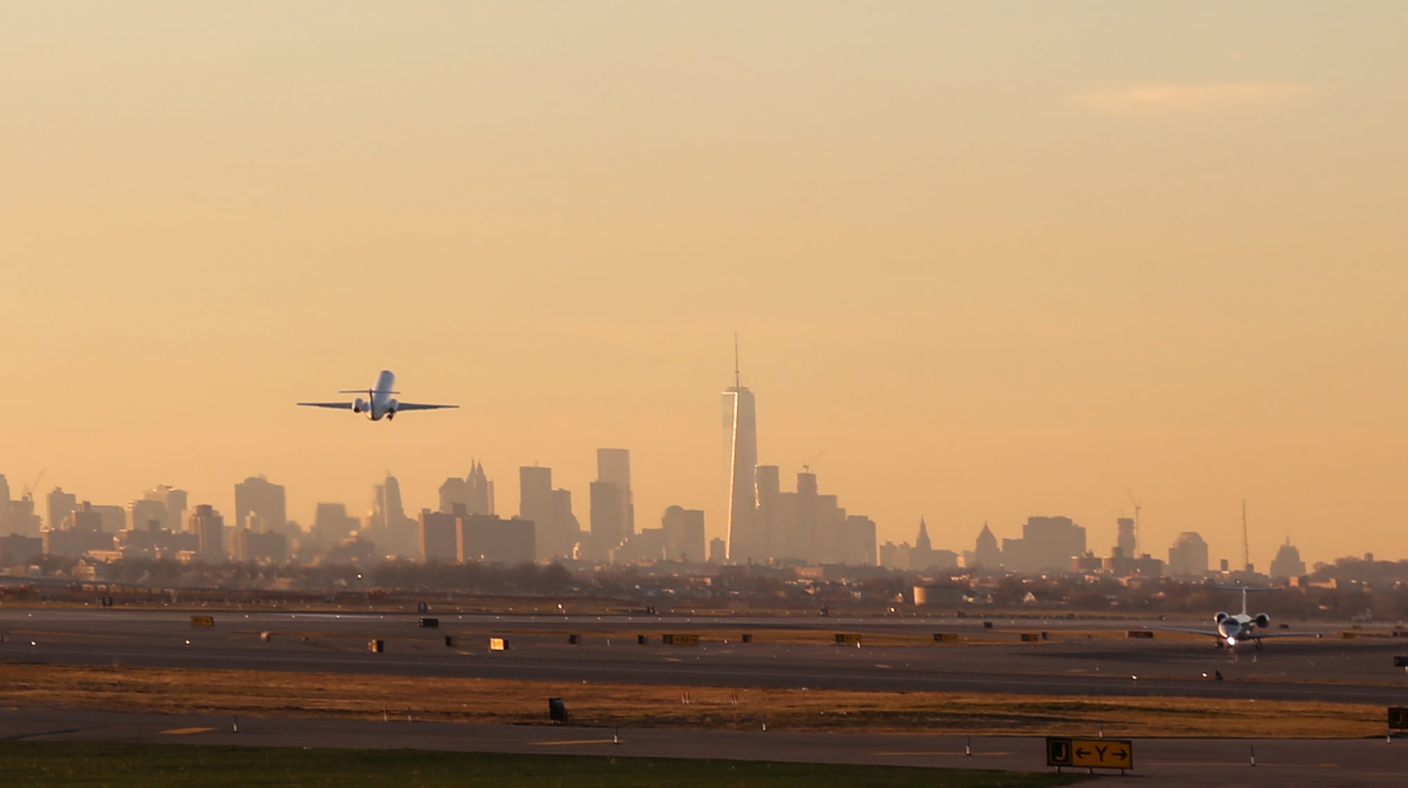 An airplane takes off from a runway at JFK Airport in Queens with NYC skyline in the background. The sun casts a warm glow, illuminating the tall buildings and creating a serene atmosphere. Another plane is visible on the runway.