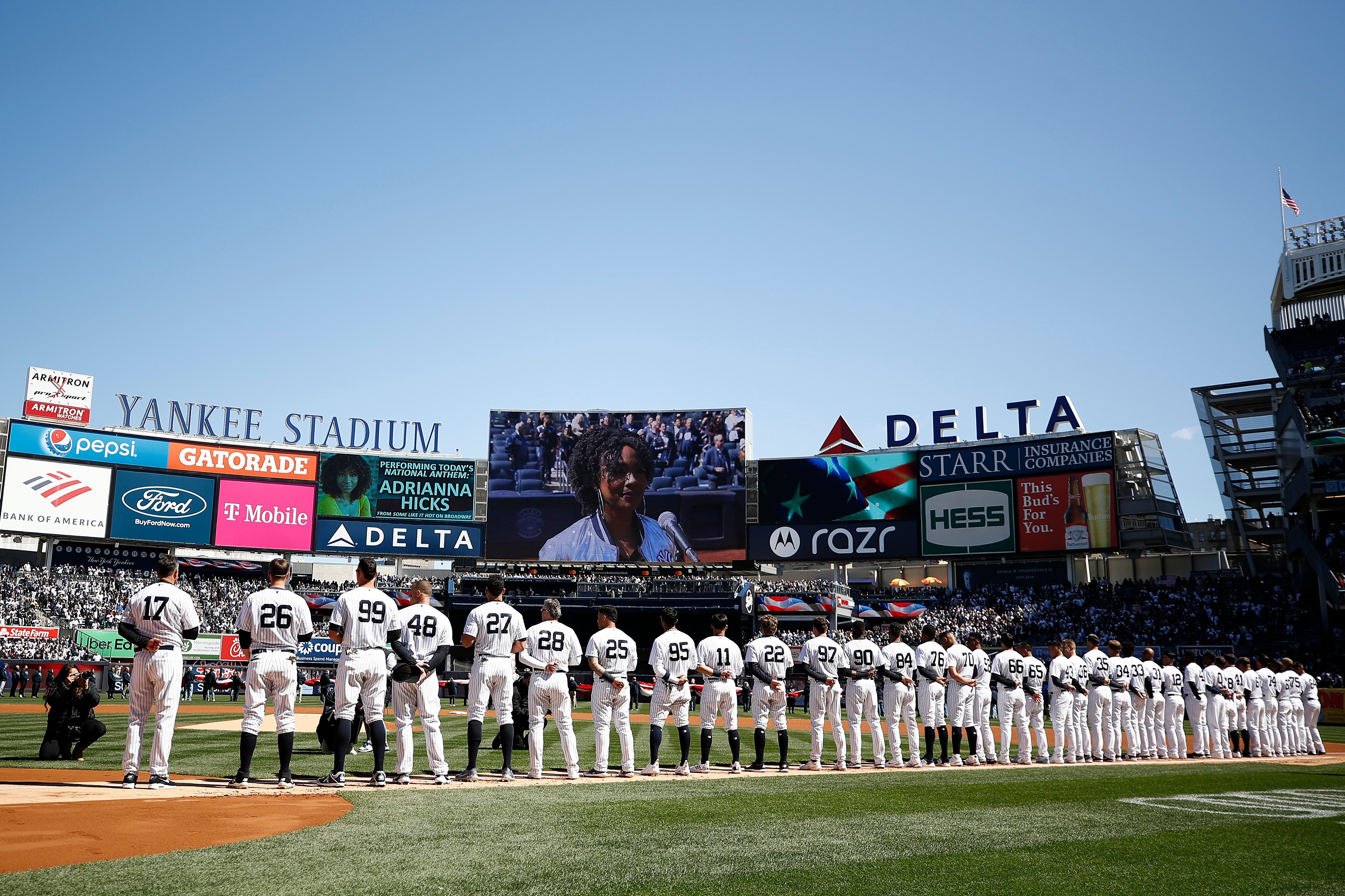A baseball team lines up on the field at Yankee Stadium for opening day, facing an American flag displayed on large screens. The players wear white uniforms with dark numbers. Advertisements for various brands are visible in the background.