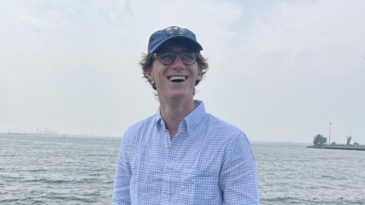 A man wearing a blue cap and glasses, Campbell Munn, smiling widely while standing near a body of water. He is dressed in a light blue, checkered long-sleeve shirt. The background shows a cloudy sky and distant shoreline.