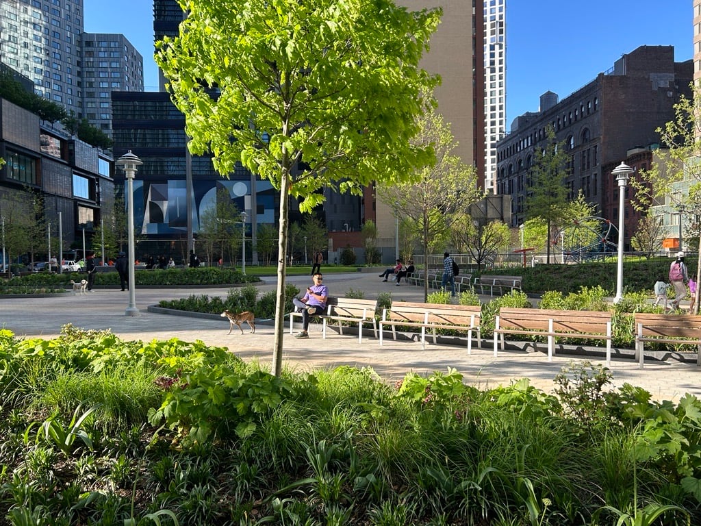 Abolitionist Place Park in Brooklyn with green trees, people sitting on benches, dogs on leash, and high-rise buildings in the background.