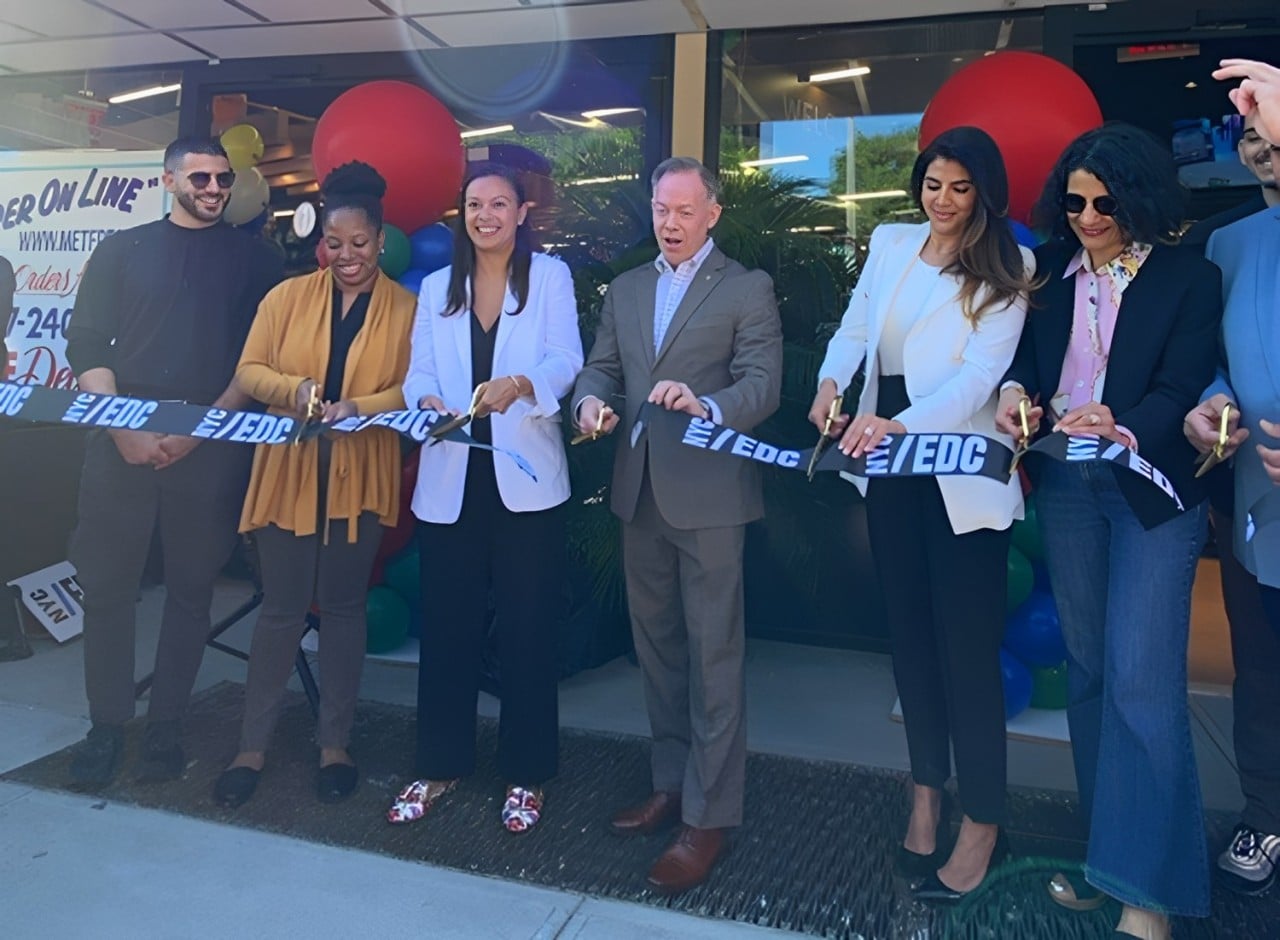 A group of six people stand in front of Met Fresh of Kensington, smiling and cutting a blue ribbon together at a ribbon-cutting ceremony. Balloons and a banner are visible in the background.